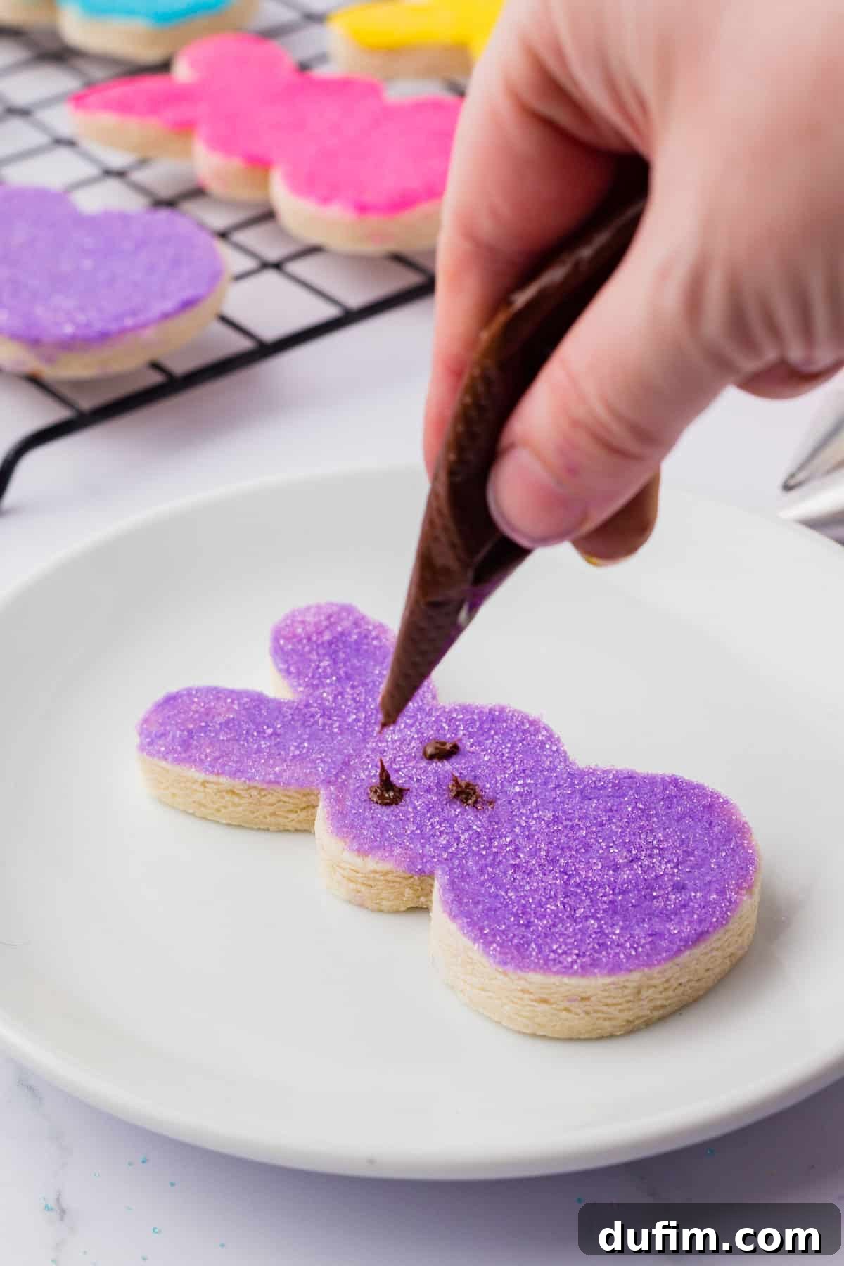 Festive Peeps Bunny Sugar Cookies 8 Peeps Bunny sugar cookie on a white plate with purple frosting and sugar on top being decorated with chocolate eyes and a nose