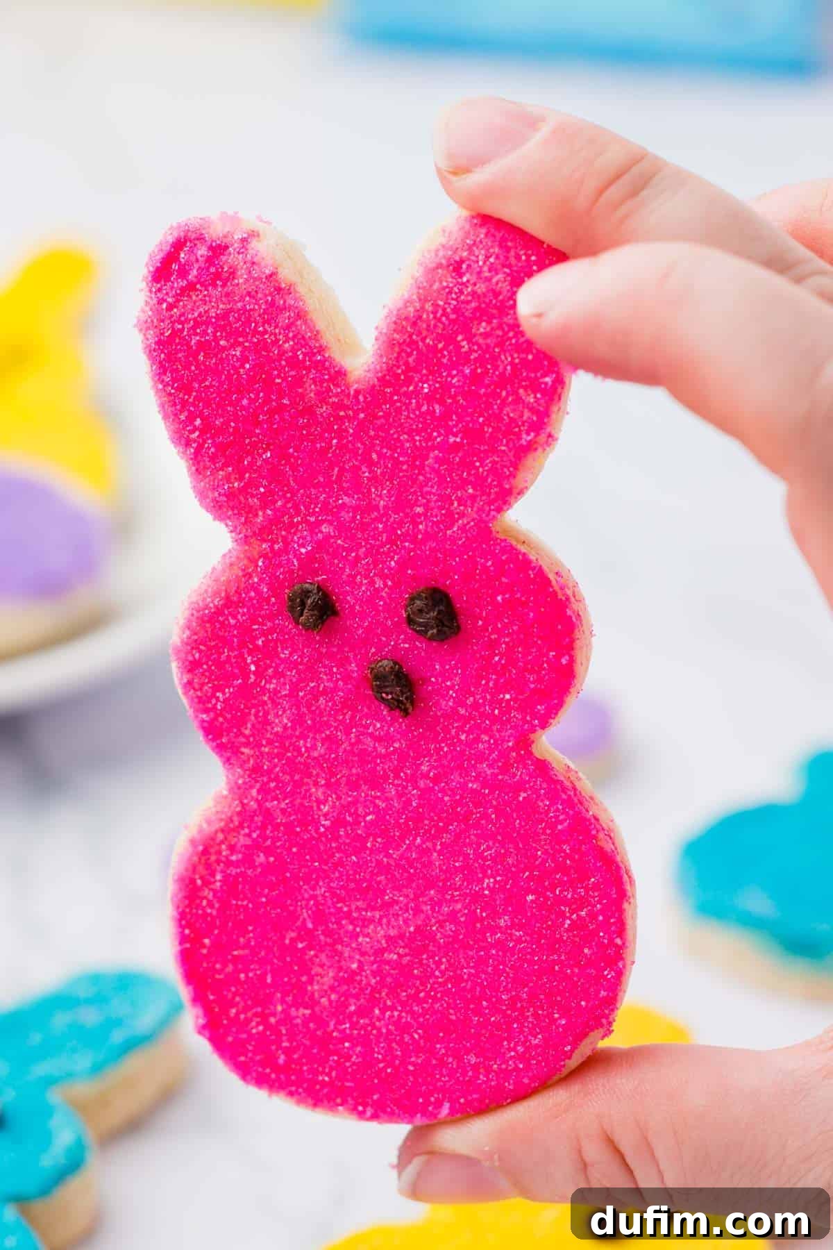 Festive Peeps Bunny Sugar Cookies 9 a pink peeps bunny sugar cookie being held up with a blurred background of other blue, yellow and purple cookies