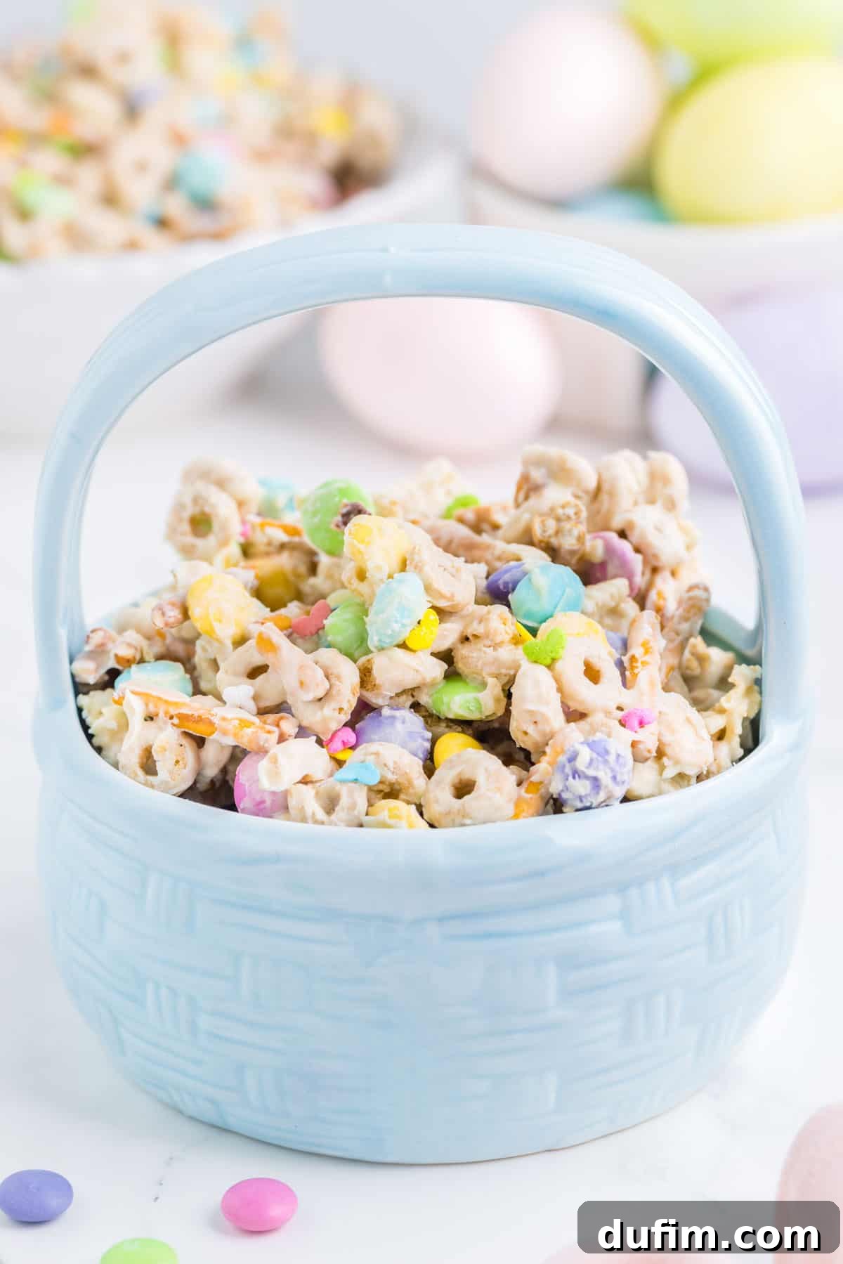 Easter bunny bait snack mix in a blue Easter basket dish with easter eggs and more bunny bait in the background and a few M&Ms around it