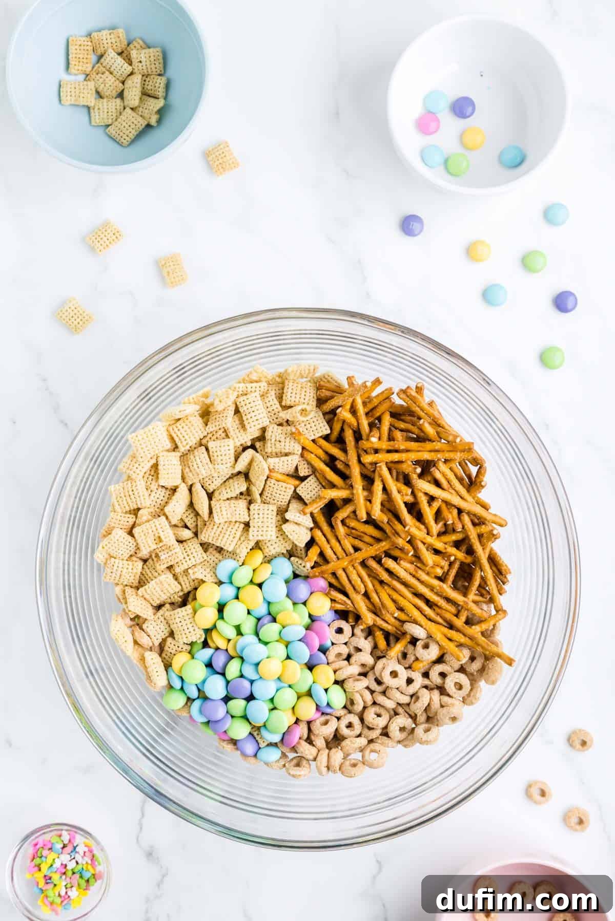 dry ingredients for bunny bait in a clear glass bowl surrounding by a bowl of sprinkles, a bowl with cheerios in it, a bowl of rice chex and a bowl of m&ms