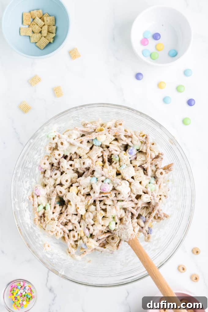 Bunny bait snack mix in a clear glass bowl surrounding by a bowl of sprinkles, a bowl with cheerios in it, a bowl of rice chex and a bowl of m&ms