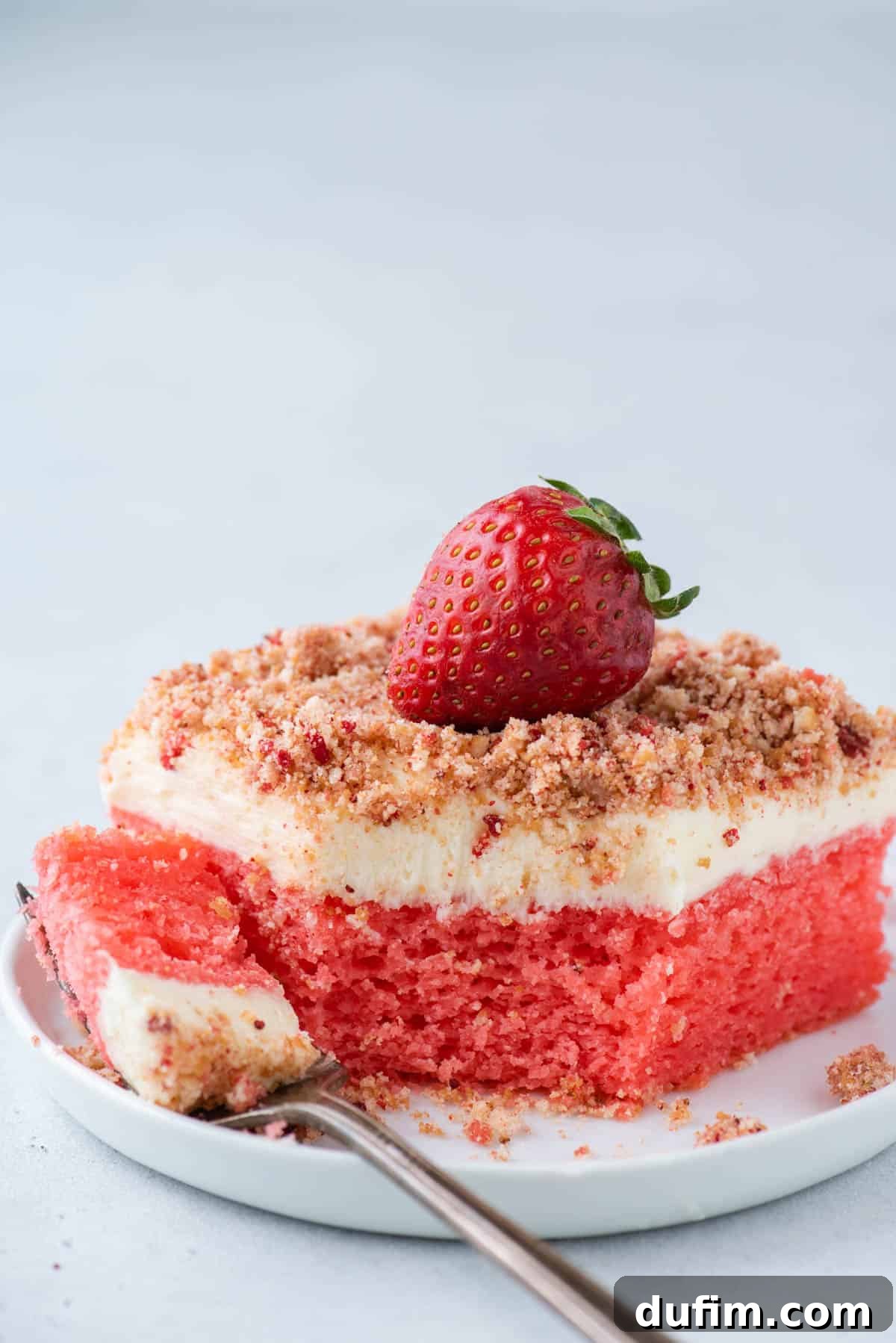 A perfectly sliced piece of Strawberry Crunch Cake on a white plate with a fork, showing the vibrant layers and crunchy topping