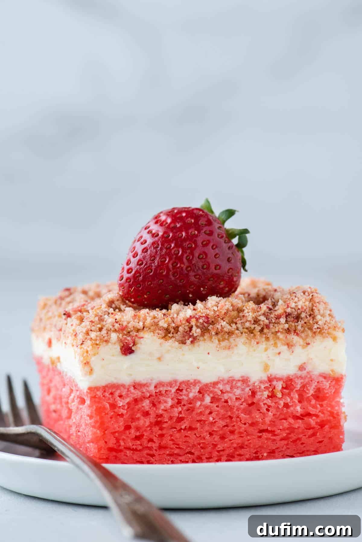 A slice of Strawberry Crunch Cake on a white plate with a fork and a whole fresh strawberry on top, showcasing the texture
