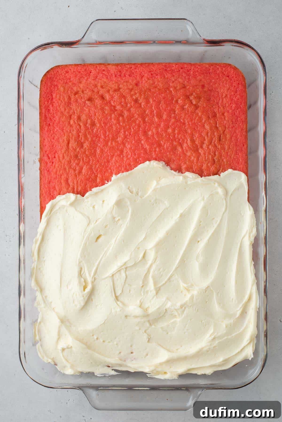 Strawberry cake in a glass baking dish, half covered in smooth vanilla frosting