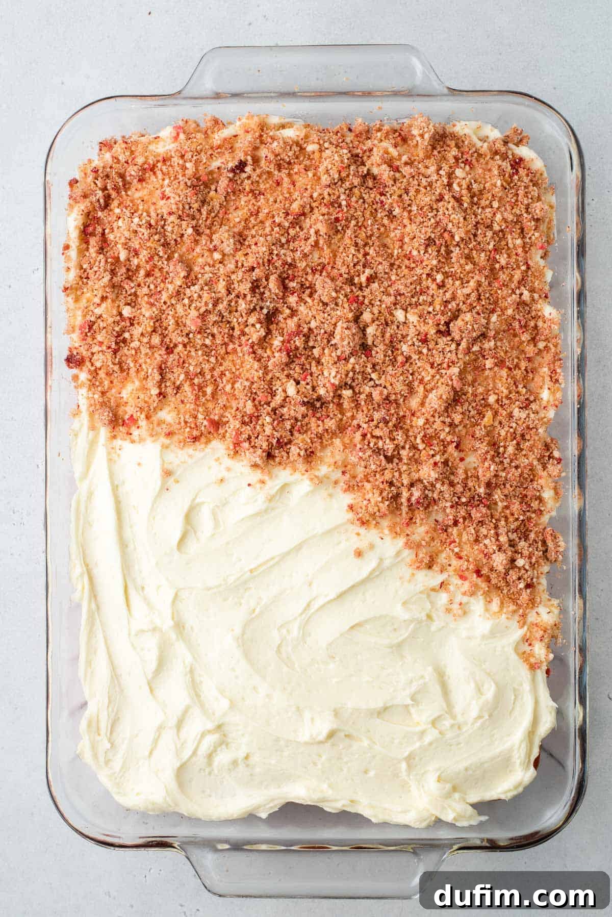 Strawberry cake in a glass baking dish, fully covered in vanilla frosting and half covered in the vibrant strawberry crunch topping