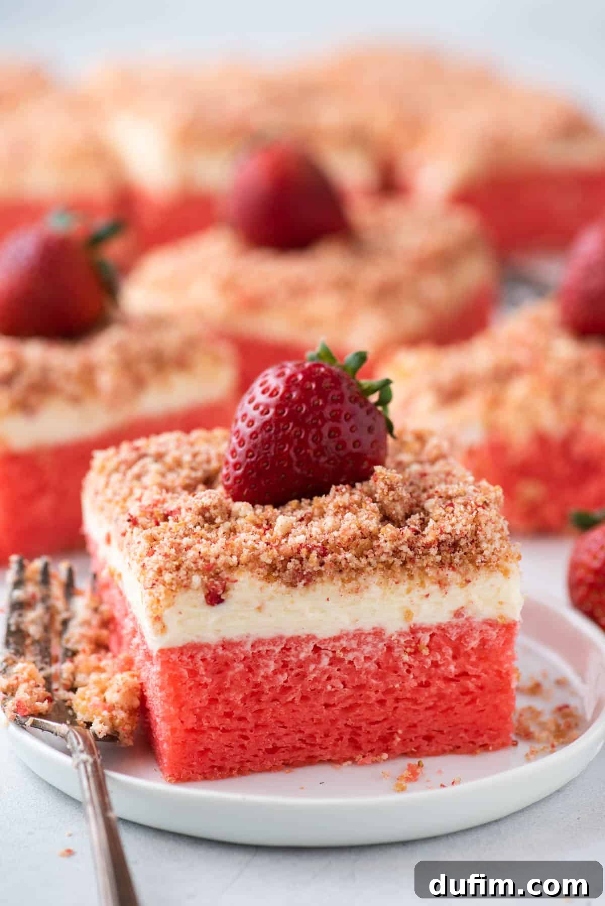 A slice of Strawberry Crunch Cake on a white plate with a fork, with more slices of the cake blurred in the background, ready to be enjoyed