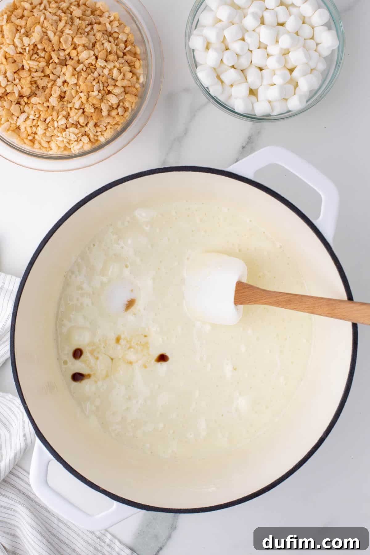 Oreo Dream Krispie Bars 6 melted mini marshmallows in a white pot surrounded by a bowl of rice krispies, a bowl of mini marshmallows and a kitchen towel