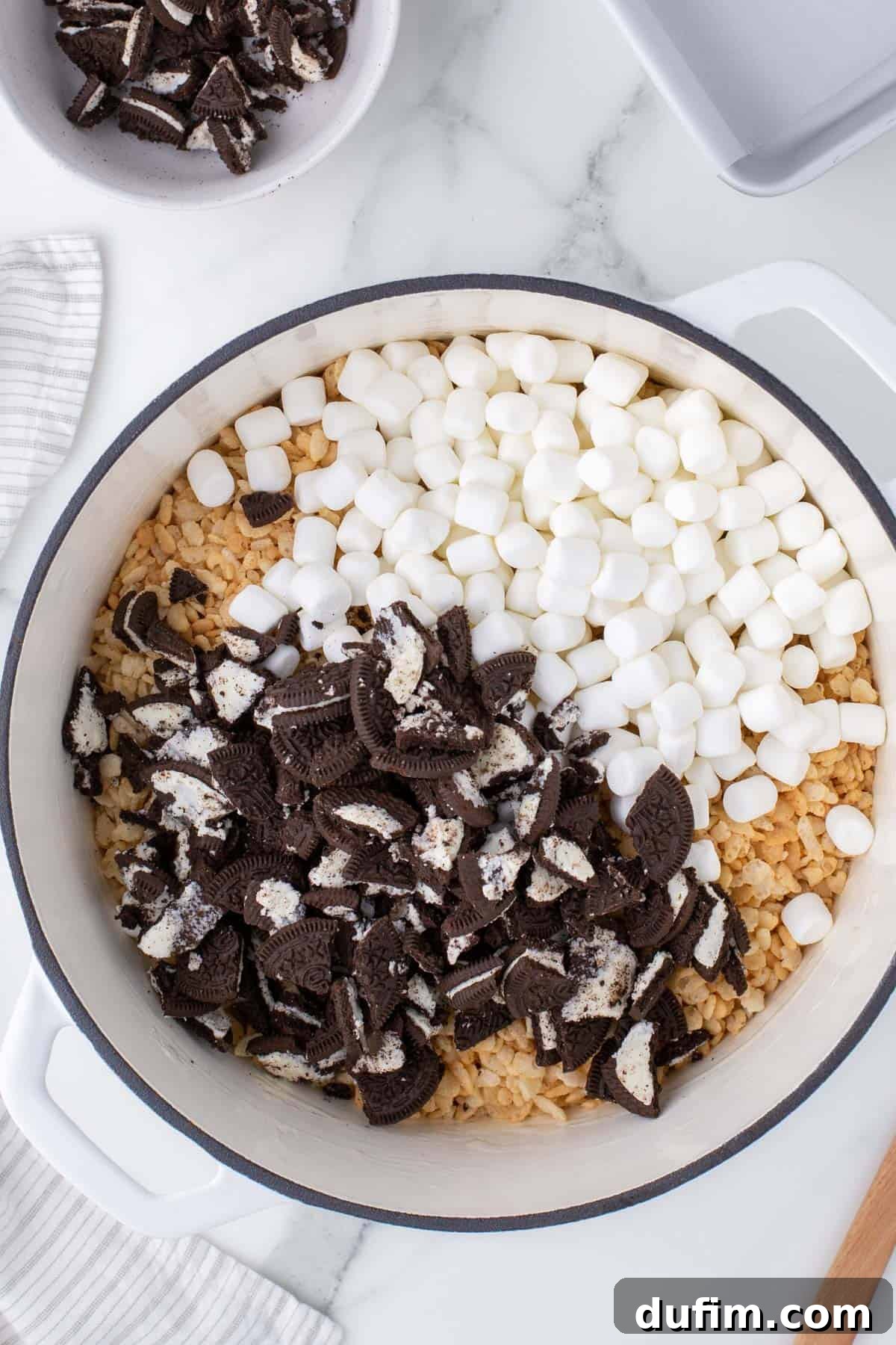 Oreo Dream Krispie Bars 7 crumbled oreo cookies, mini marshmallows and rice krispie cereal in a white pot surrounded by a kitchen town, wooden spatula and a bowl of more crumbled oreos.
