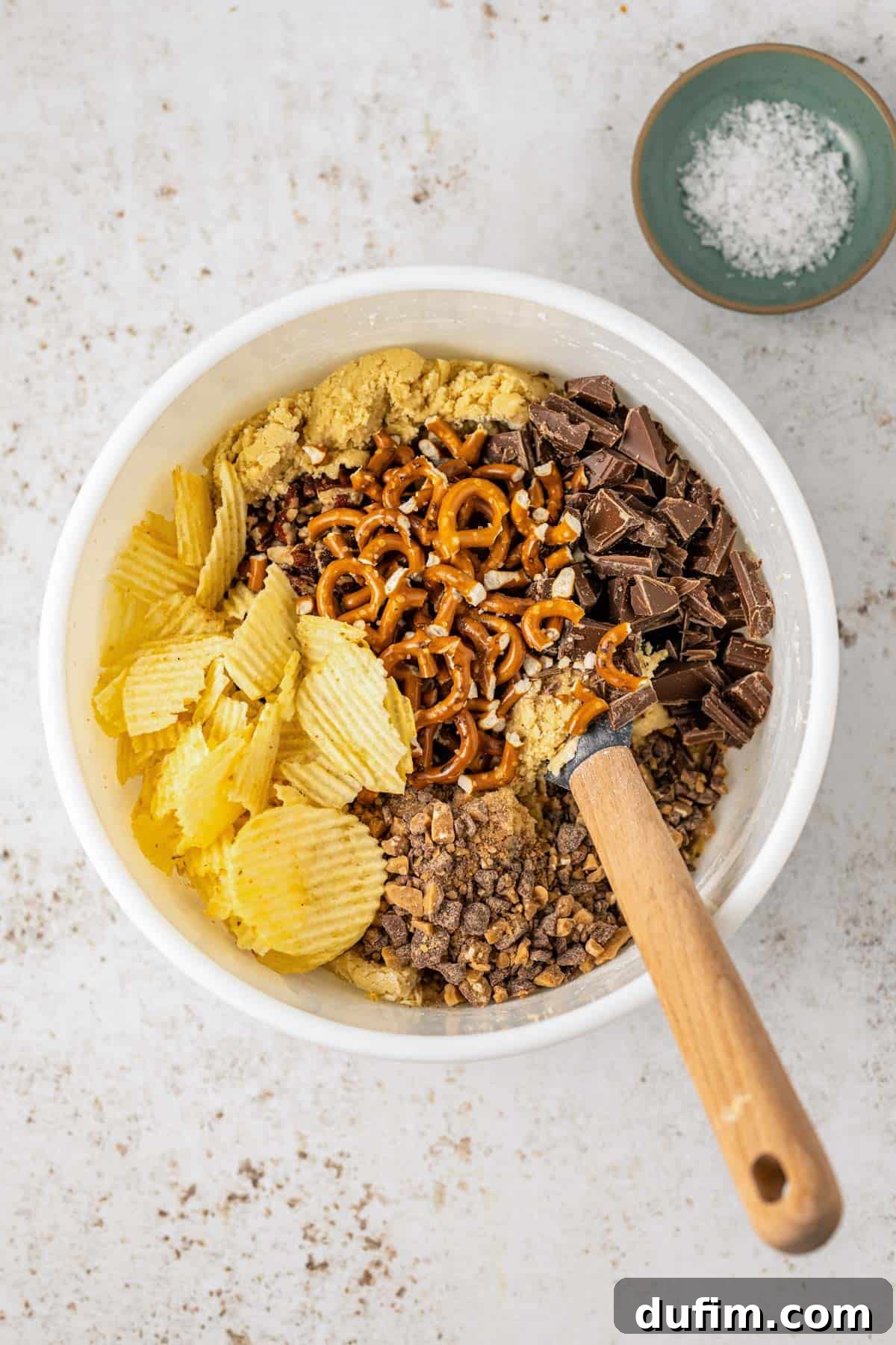 A white bowl brimming with unbaked Kitchen Sink Cookie dough, topped with generous piles of potato chips, toffee bits, pretzels, and chocolate chunks, with a wooden spatula resting inside the bowl and a small dish of flaky salt on the side