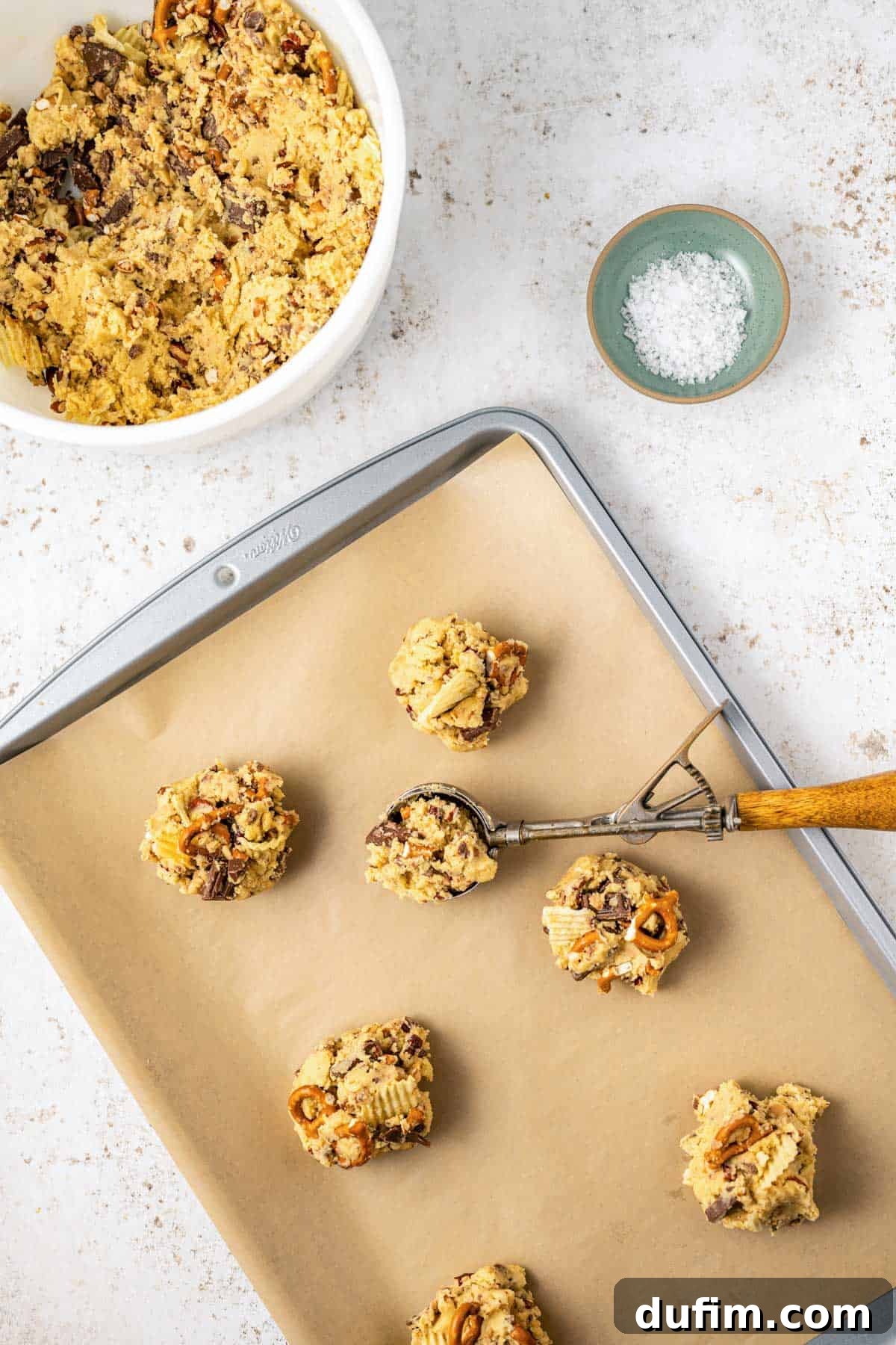 A baking sheet lined with parchment paper, holding perfectly portioned Kitchen Sink Cookie dough balls, with one still in the cookie scoop, a small bowl of salt nearby, and a larger bowl of remaining cookie dough beside it