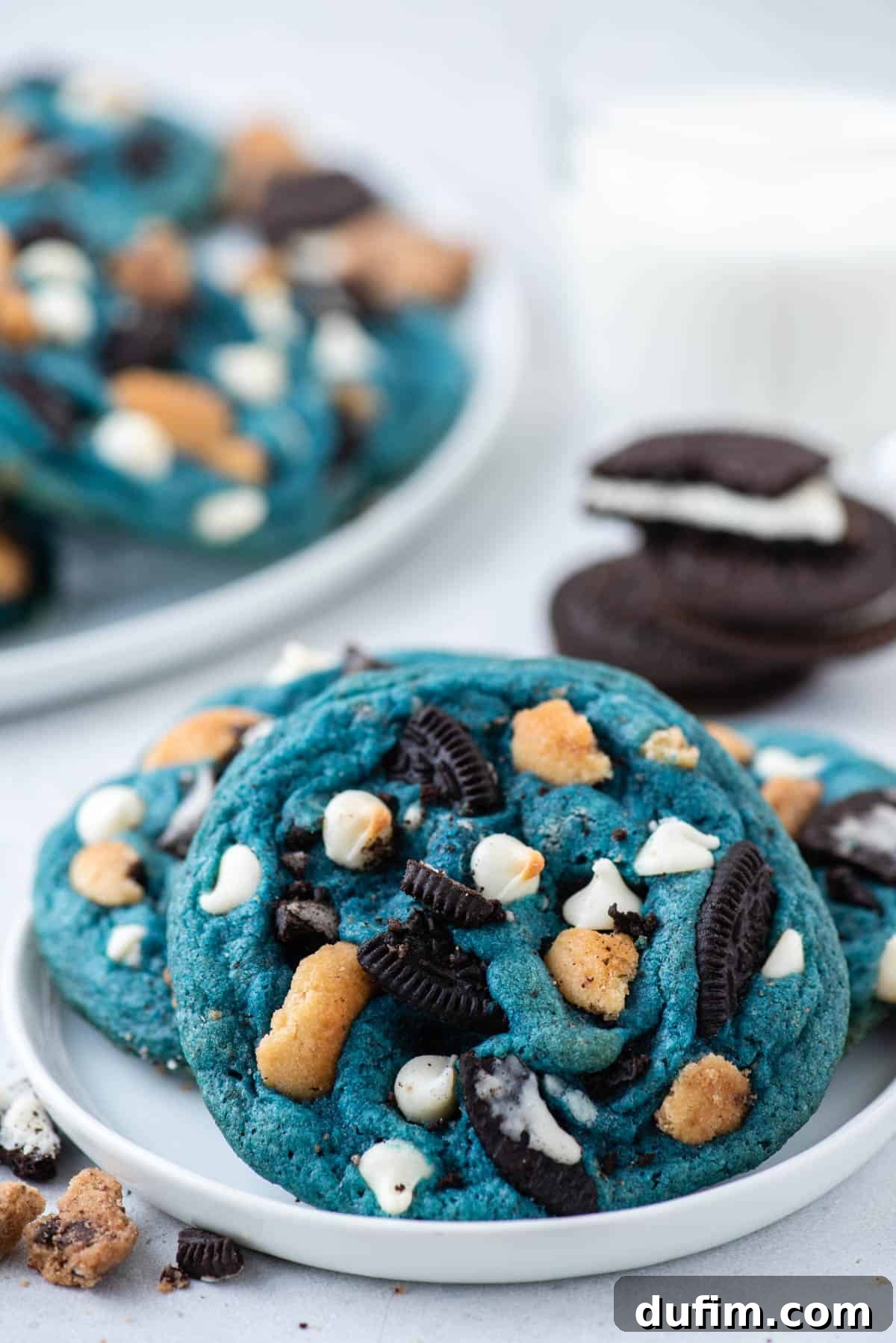 A white plate with three vibrant blue Cookie Monster Cookies on it, surrounded by cookie crumbs. Oreos and additional cookies are blurred in the background, hinting at the delicious ingredients.