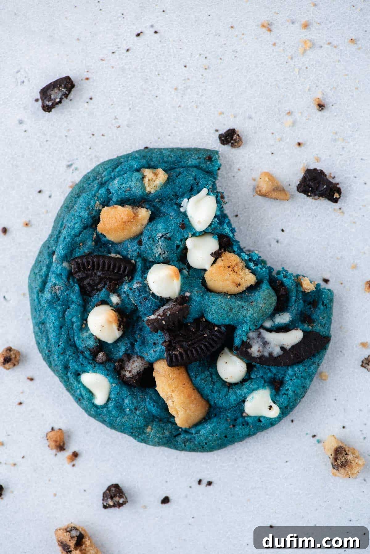 A single vibrant blue Cookie Monster Cookie with a bite taken out of it, revealing the white chocolate chips, Chips Ahoy, and Oreo pieces inside. Cookie crumbs are scattered around the plate.