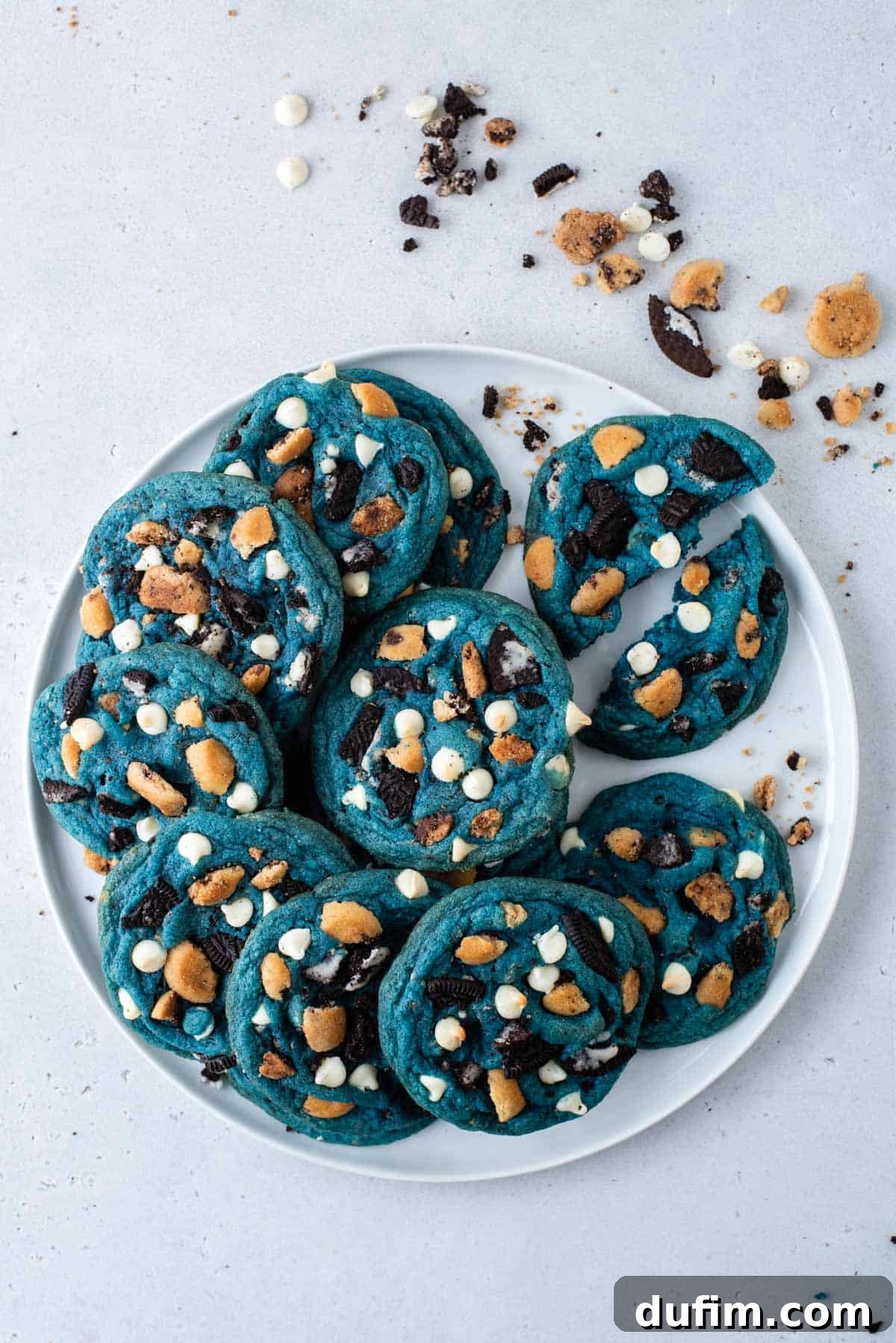 A ceramic plate filled with vibrant blue Cookie Monster Cookies. One cookie on the right is broken in half, revealing its soft, chewy interior studded with white chocolate chips, Oreo, and Chips Ahoy pieces. Cookie crumbs are artfully sprinkled around.