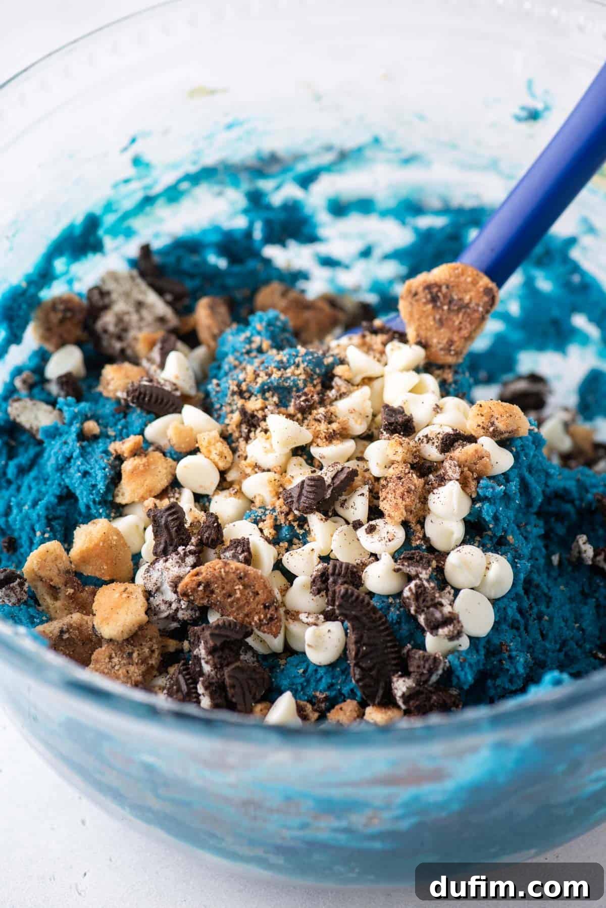 A close-up view of a bowl of vibrant blue cookie monster cookie batter, generously topped with white chocolate chips, chunky Oreo pieces, and broken Chips Ahoy cookie bits, ready to be mixed in.