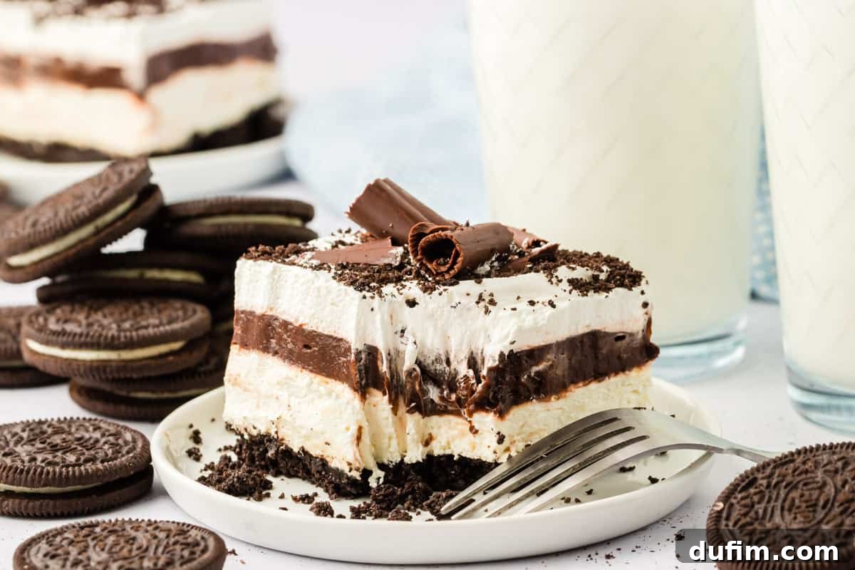 Ultimate Chocolate Lasagna Indulgence 2 A perfectly sliced chocolate lasagna on a white plate with a fork, showing a bite taken out. It's surrounded by whole Oreo cookies, cookie crumbs, and a glass of milk in the background.
