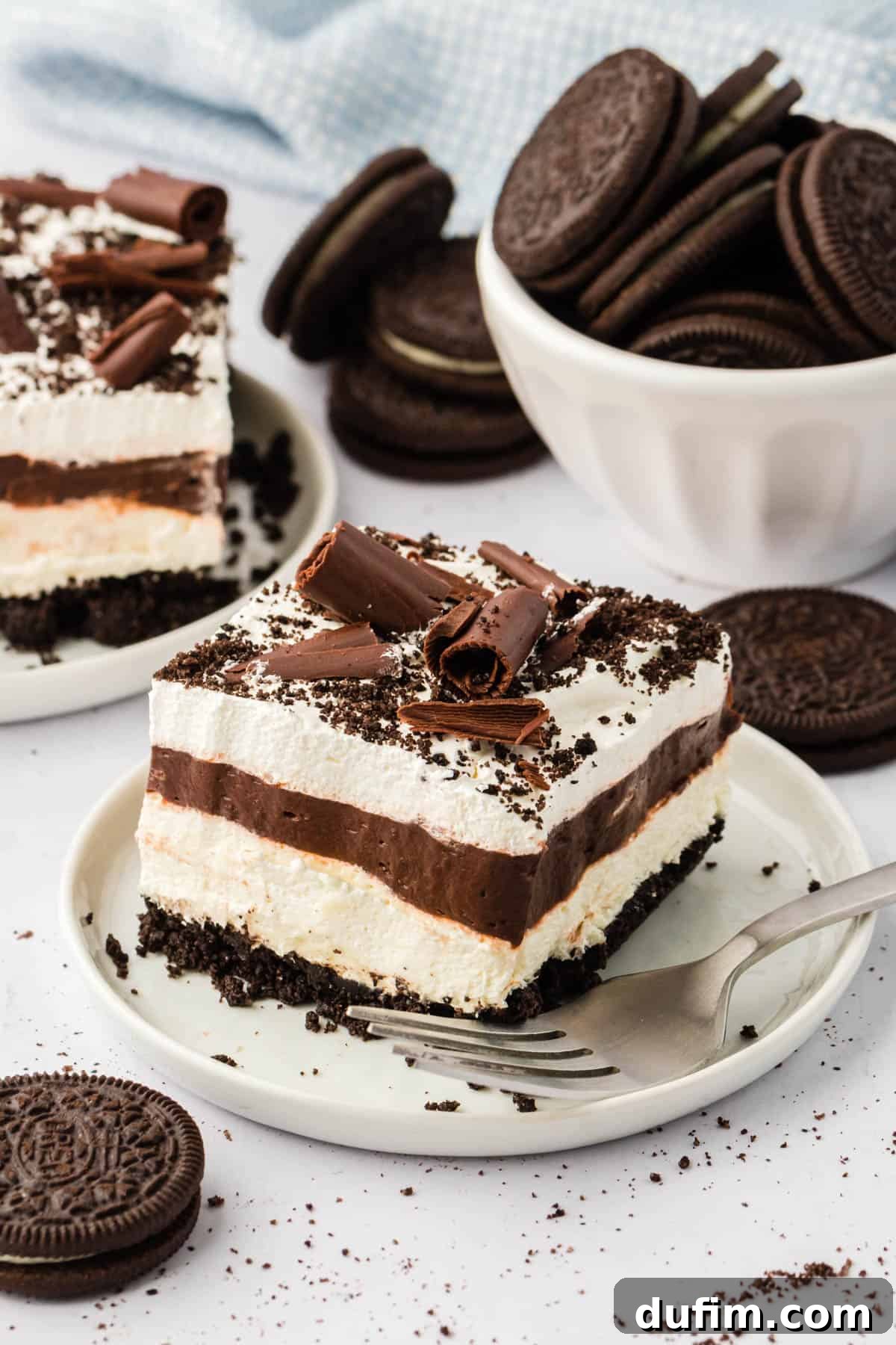Ultimate Chocolate Lasagna Indulgence 6 A luscious slice of chocolate lasagna on a white plate, with a fork mark from a bite, surrounded by whole Oreos, crumbs, and another slice in the background.