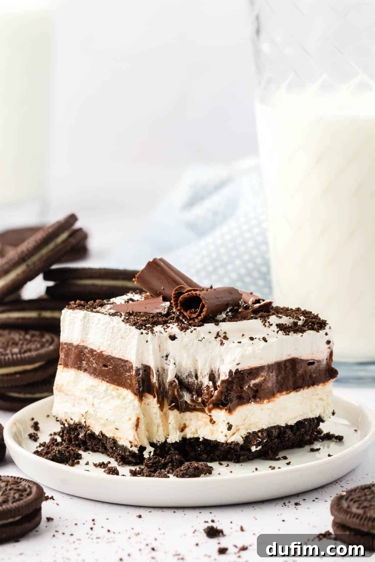 Ultimate Chocolate Lasagna Indulgence 7 A close-up view of a slice of chocolate lasagna on a white plate with a bite taken, surrounded by whole Oreo cookies, cookie crumbs, and a refreshing glass of milk in the background.