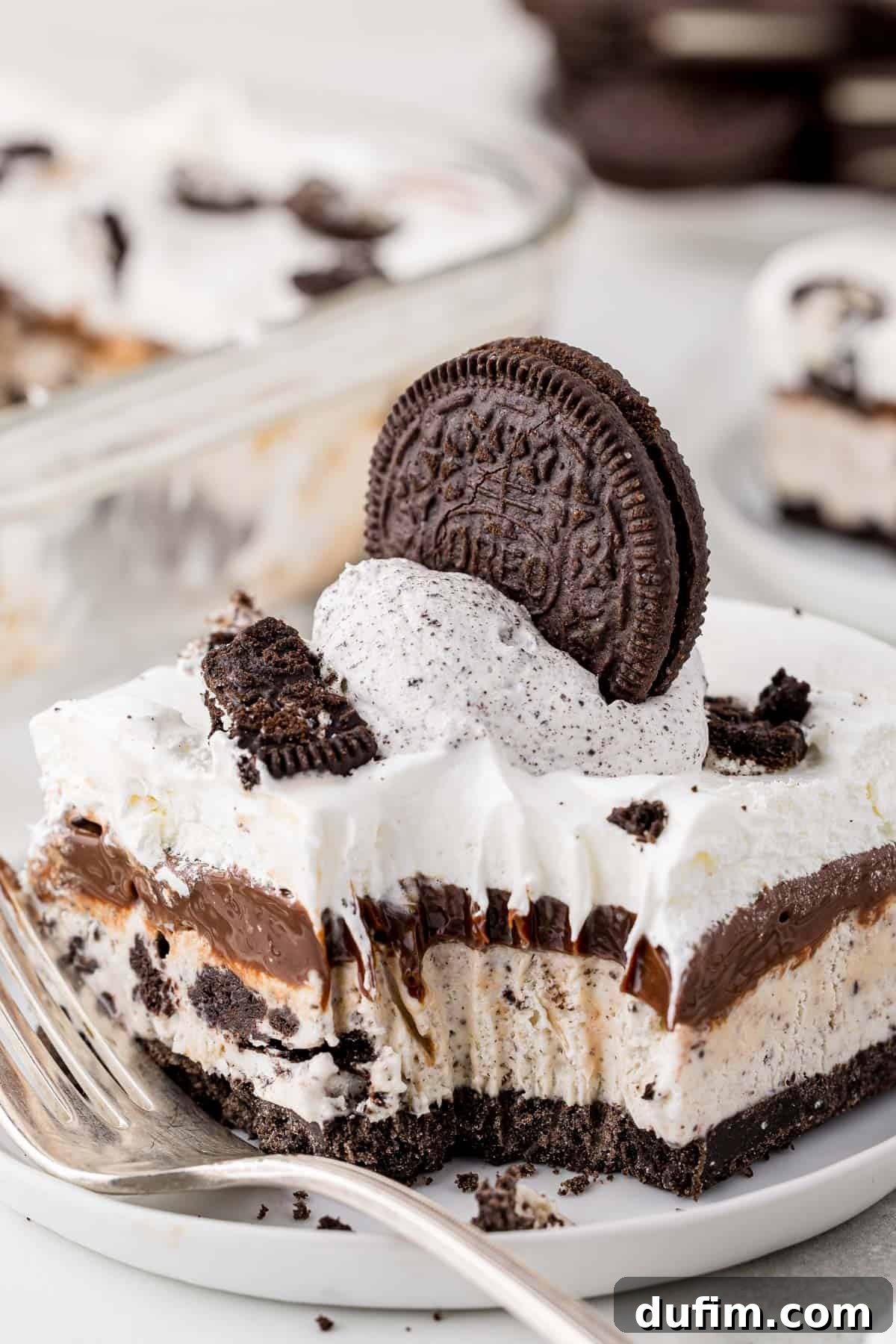 A perfectly sliced piece of Oreo ice cream cake on a white plate, with a bite taken out and a fork resting beside it, showcasing the distinct layers of cookie crust, ice cream, fudge, and whipped topping.
