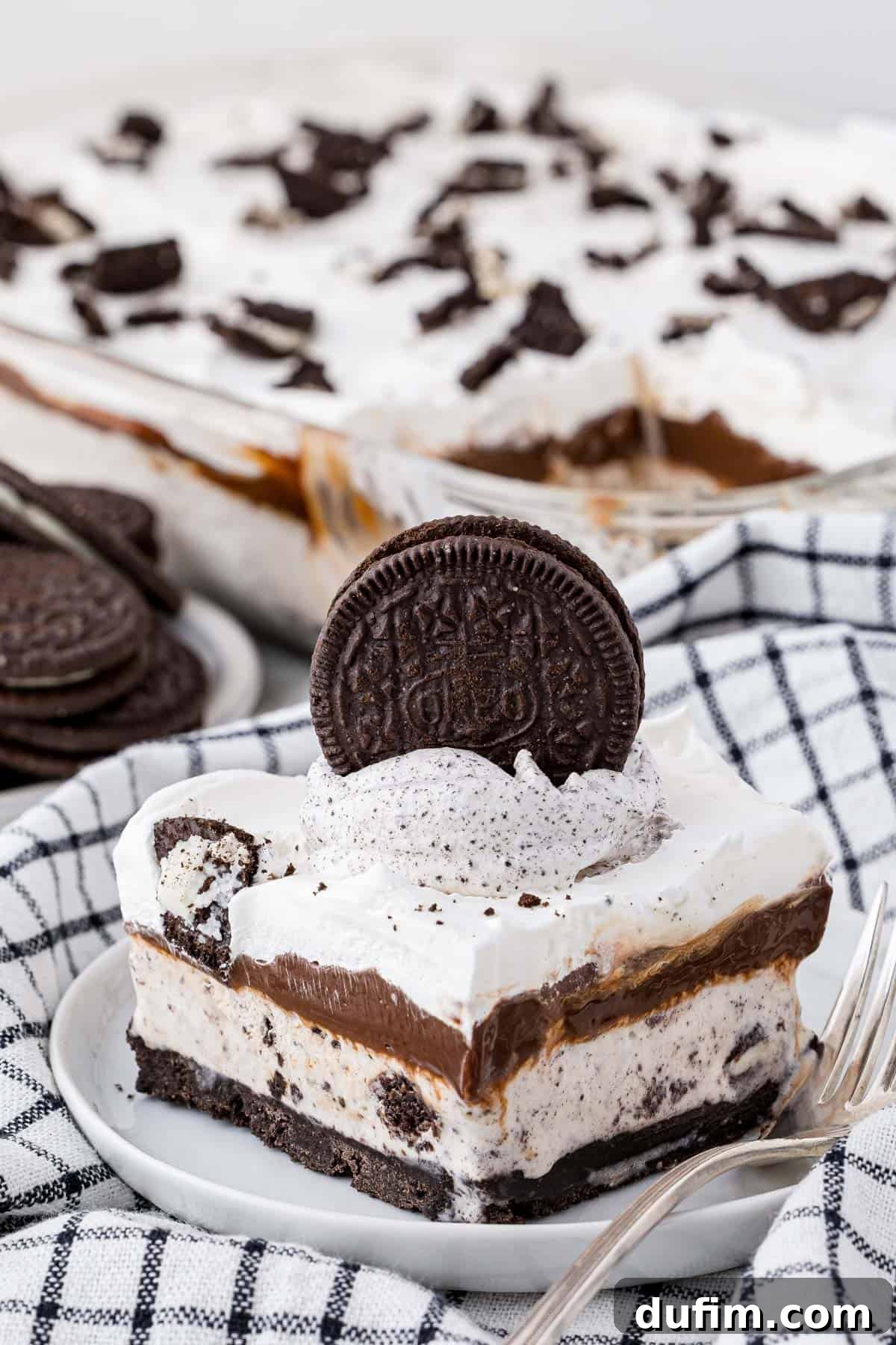 A beautiful slice of homemade Oreo ice cream cake served on a white plate with a fork, resting on a black and white checkered kitchen towel. More ice cream cake and whole Oreos are visible in the background.