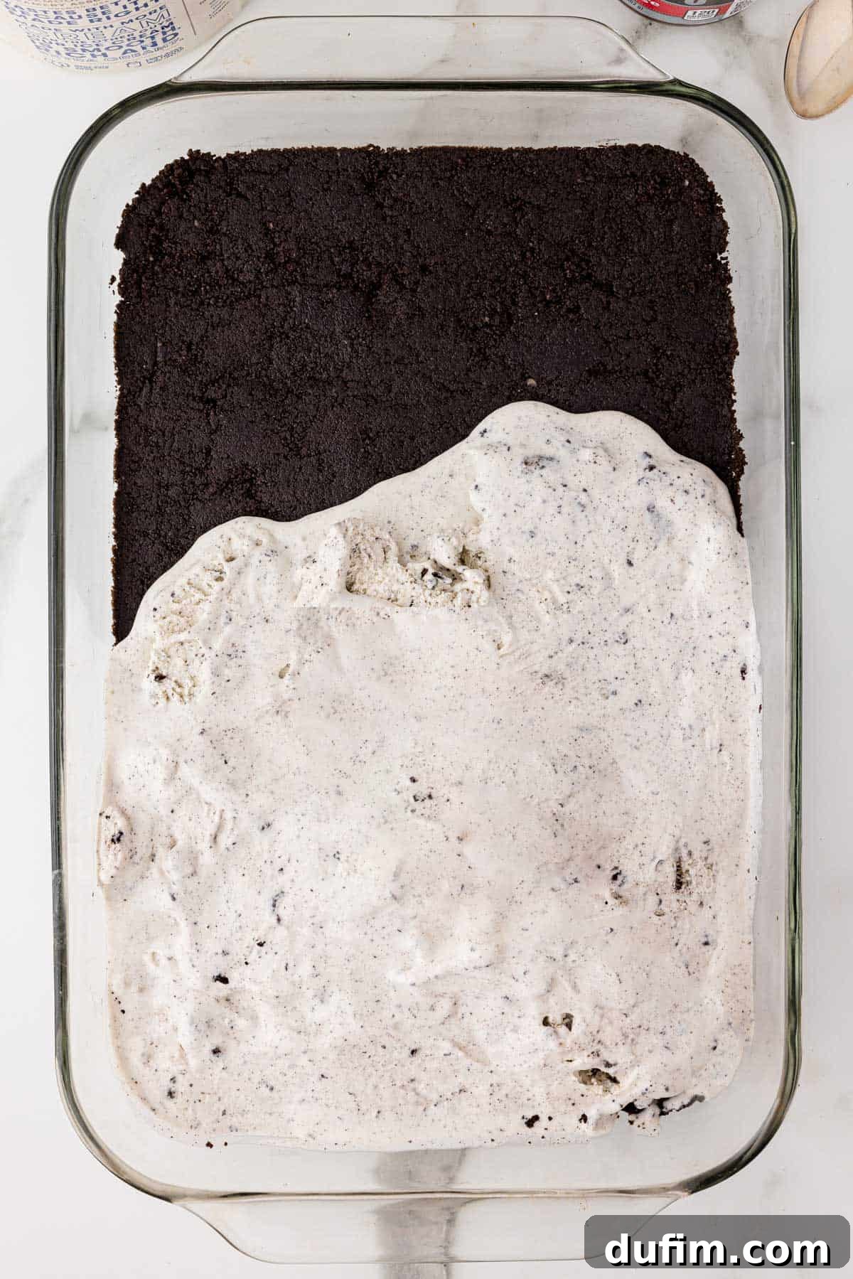 The initial layers of Oreo ice cream cake being assembled in a baking dish, showing the dense Oreo cookie crust at the bottom and a partially spread layer of softened cookies and cream ice cream on top.
