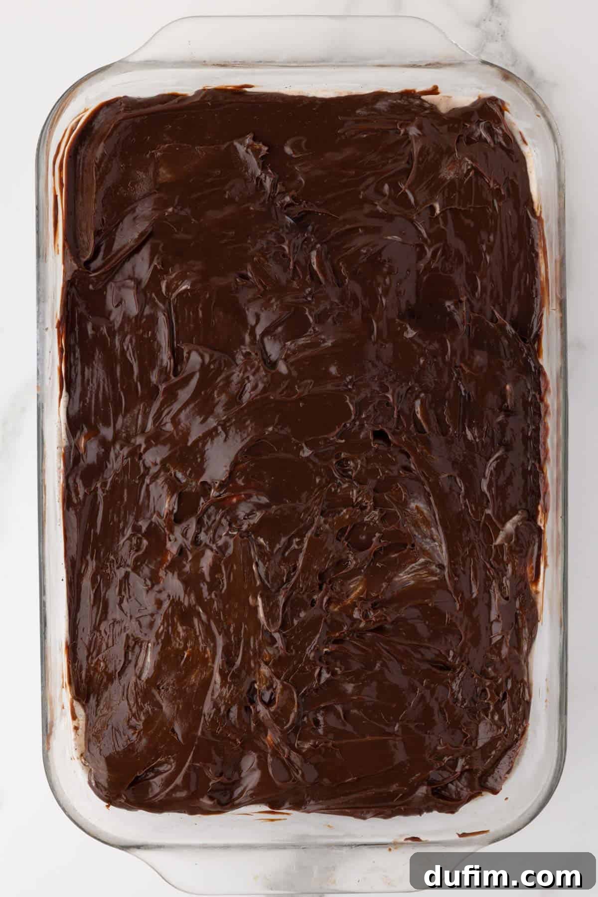 The second stage of layering an Oreo ice cream cake, with the hot fudge layer spread smoothly over the frozen ice cream layer, ready for the next step.