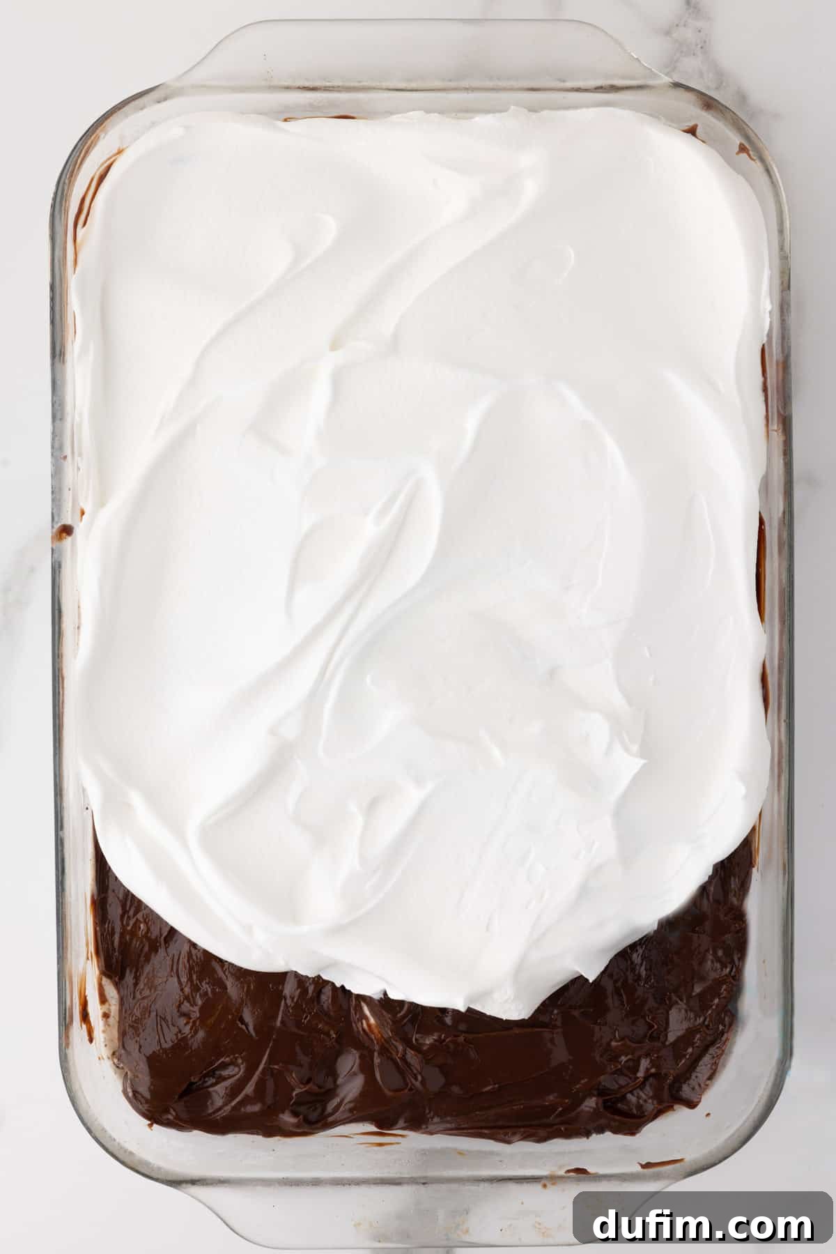 The third layer of Oreo ice cream cake being applied, showing the hot fudge layer covered halfway with fluffy cool whip, demonstrating the smooth spreading process.