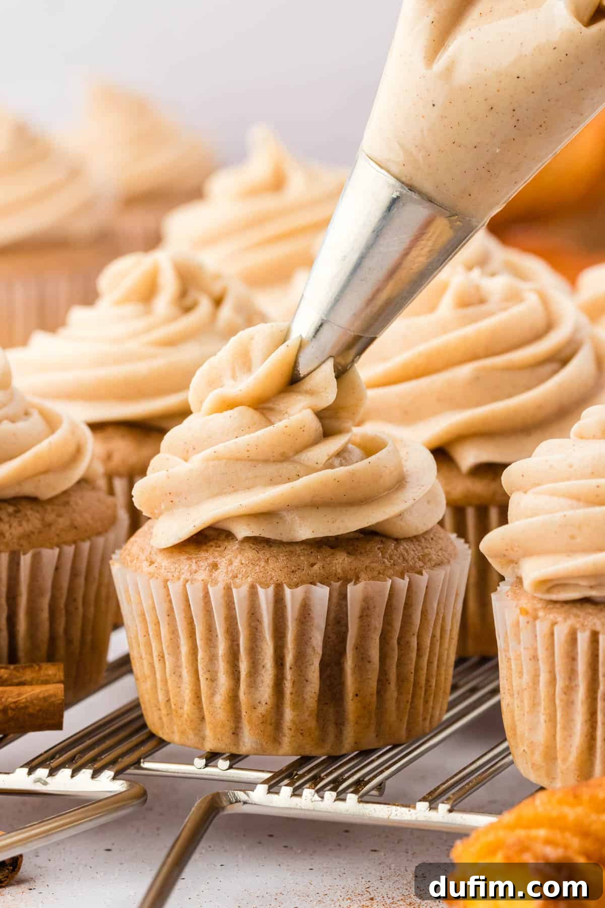 Dreamy Cinnamon Cream Cheese Frosting 3 Cinnamon cream cheese frosting being expertly piped onto a freshly baked cupcake resting on a wire cooling rack, with other iced cupcakes visible in the background, showcasing the ease of application.