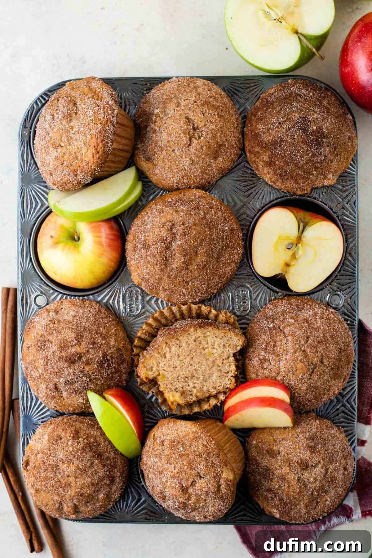 Tender Apple Cinnamon Treats 10 A stunning arrangement showcasing a muffin pan filled with golden-brown apple cinnamon muffins, fresh red and green apple slices, half apples, and whole apples, all beautifully complemented by fragrant cinnamon sticks and more apples in the background. It truly embodies the abundance of autumn flavors.