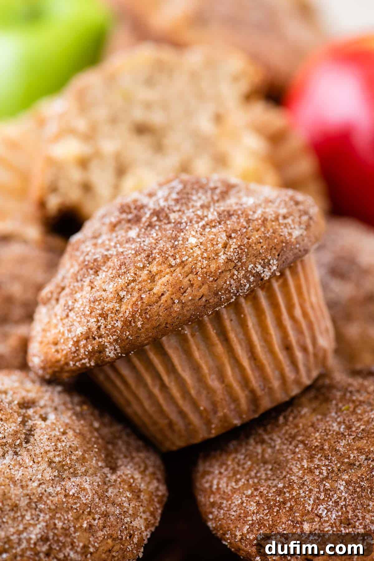Tender Apple Cinnamon Treats 11 A delectable pile of apple cinnamon muffins, generously topped with glistening cinnamon sugar, beckons with its inviting aroma. In the softly blurred background, an assortment of fresh red and green apples hints at the wholesome ingredients within.