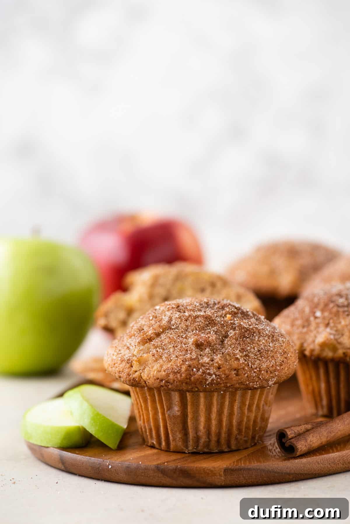 Tender Apple Cinnamon Treats 3 A beautiful display of golden-brown apple cinnamon muffins on a wooden platter, surrounded by aromatic cinnamon sticks, vibrant green apple slices, and an abundance of fresh apples in the background, showcasing the bakery-style appeal.