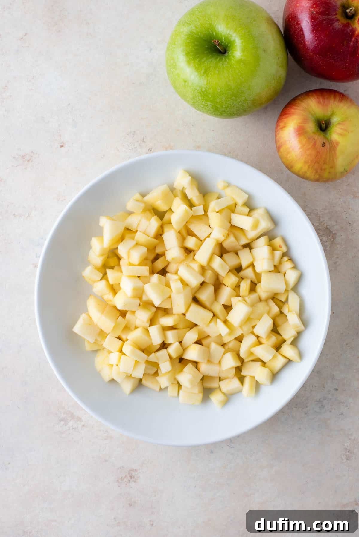 Tender Apple Cinnamon Treats 4 A pristine white bowl brimming with perfectly diced apples, flanked by three whole apples: a crisp green Granny Smith, a large vibrant red apple, and a smaller red and yellowish apple, ready for baking.