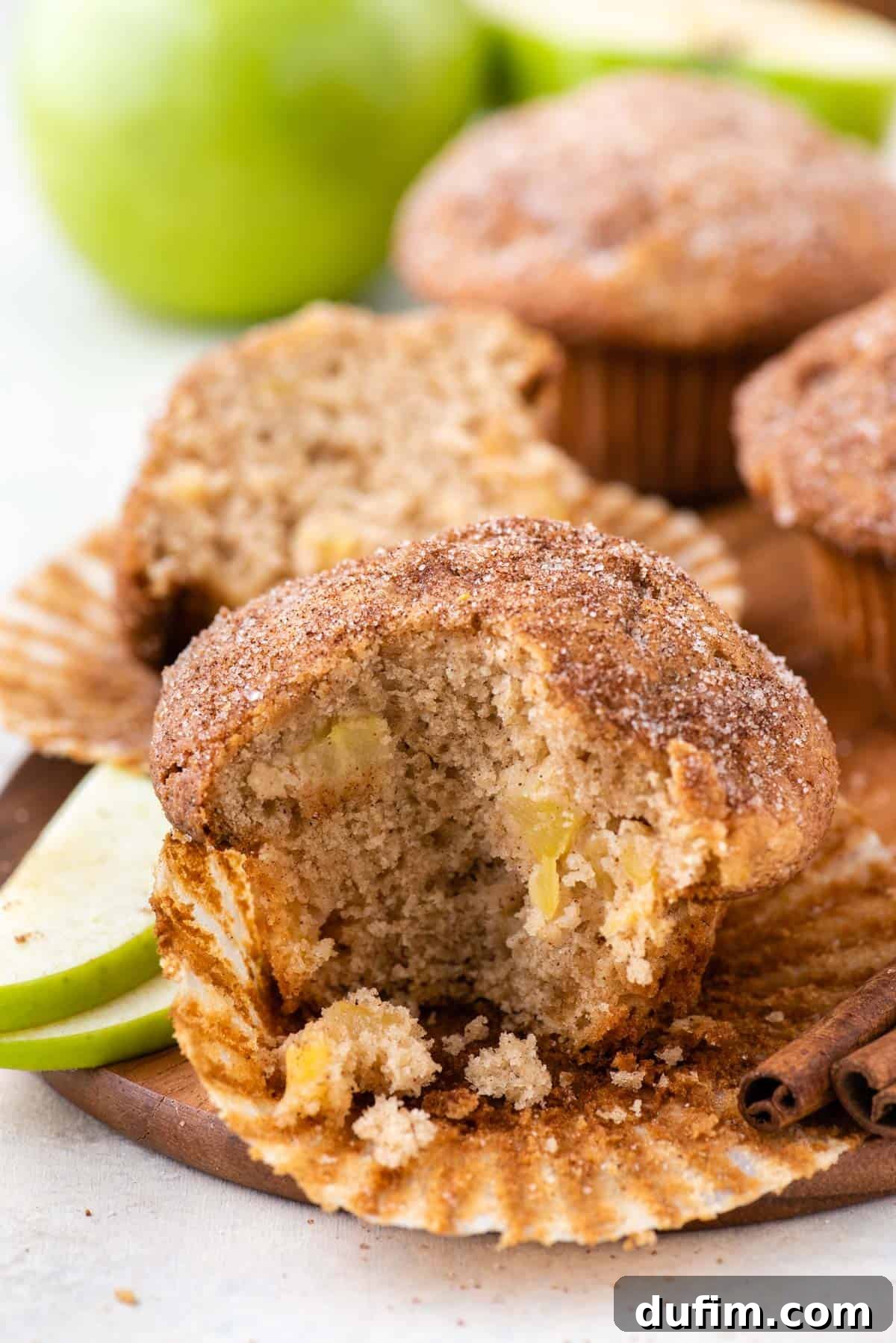 Tender Apple Cinnamon Treats 5 A partially eaten apple cinnamon muffin, still nestled in its paper liner, rests on a white plate. It's surrounded by fragrant cinnamon sticks and vibrant green apple slices, inviting a closer look at its delicious interior.