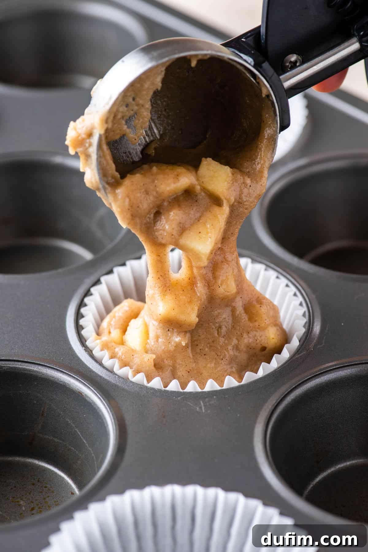 Tender Apple Cinnamon Treats 6 Close-up of apple cinnamon muffin batter being carefully scooped into a muffin liner, nestled within a muffin pan, showing the rich texture of the uncooked dough.
