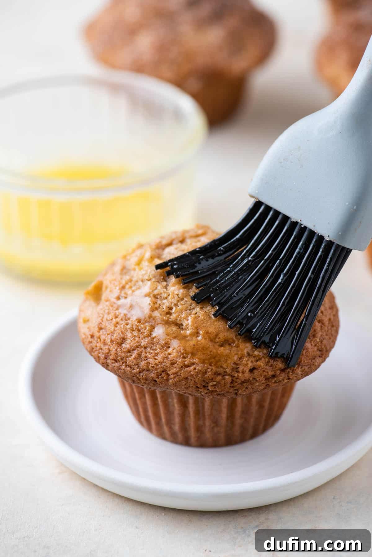 Tender Apple Cinnamon Treats 8 An apple cinnamon muffin on a white plate, getting a generous brush of melted butter with a pastry brush, with a small dish of melted butter blurred in the background, preparing for its cinnamon sugar coating.