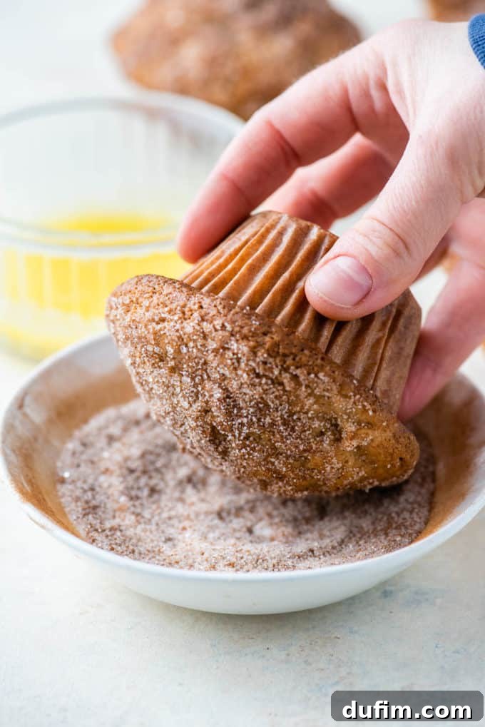 Tender Apple Cinnamon Treats 9 The top of an apple cinnamon muffin being delicately dipped into a bowl of sparkling cinnamon sugar, ready to be coated, with a dish of melted butter visible in the background.