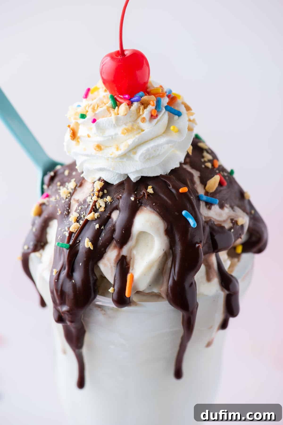 close up of a hot fudge sundae topped with sprinkles, peanuts, whipped cream and a cherry on top with a blue spoon stuck into the side