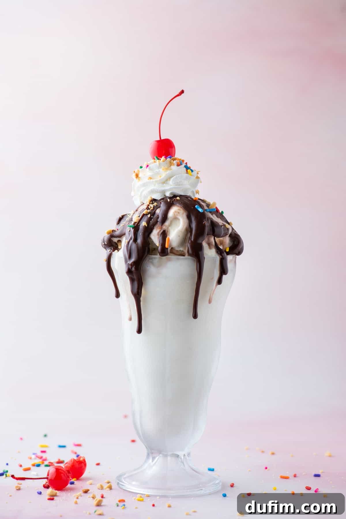 a hot fudge sunday in a tall glass surrounded by sprinkles, bits of peanuts and a couple maraschino cherries