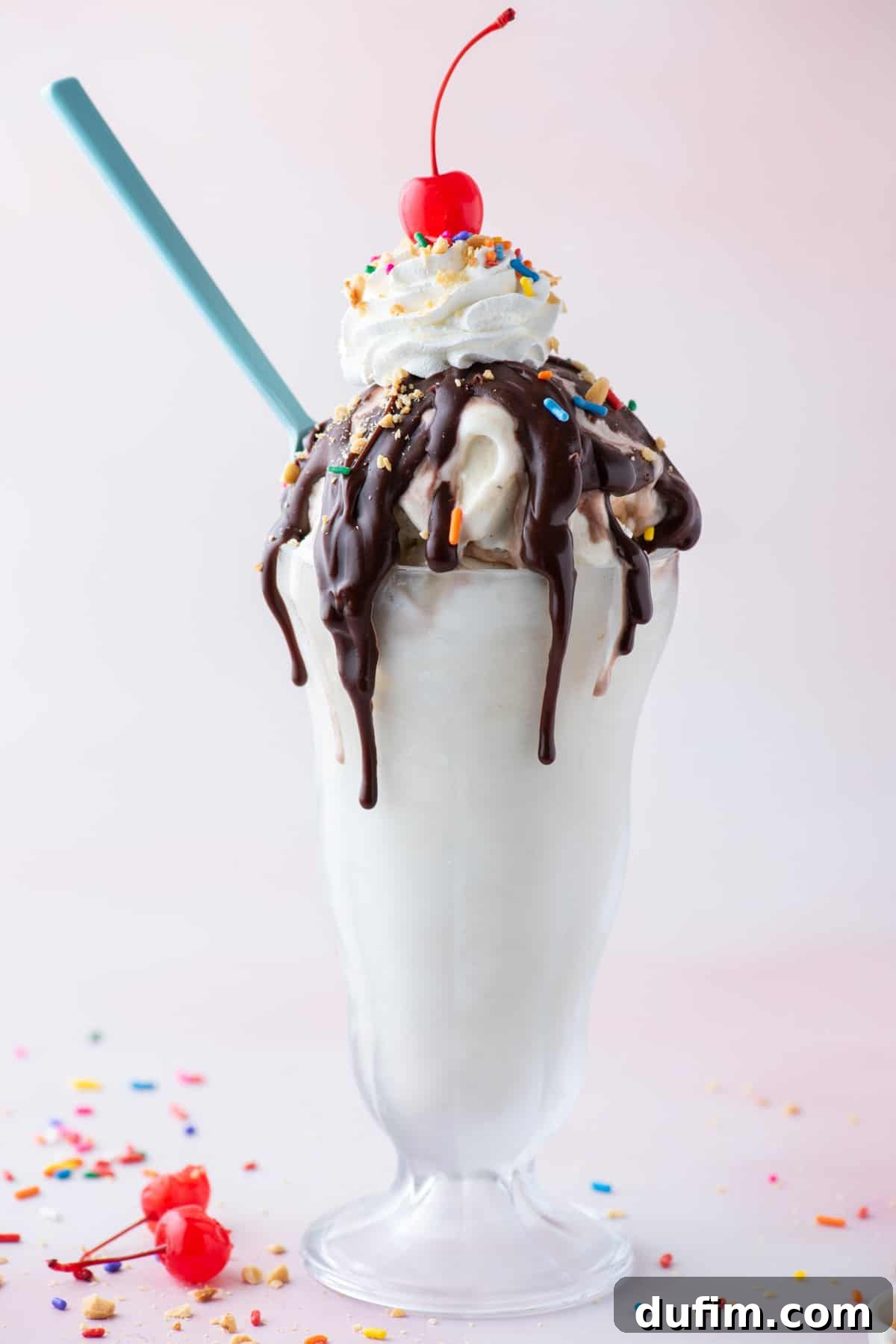 a hot fudge sundae in a tall glass surrounded by sprinkles, bits of peanuts and a couple maraschino cherries with a blue spoon stuck into the side of the sundae