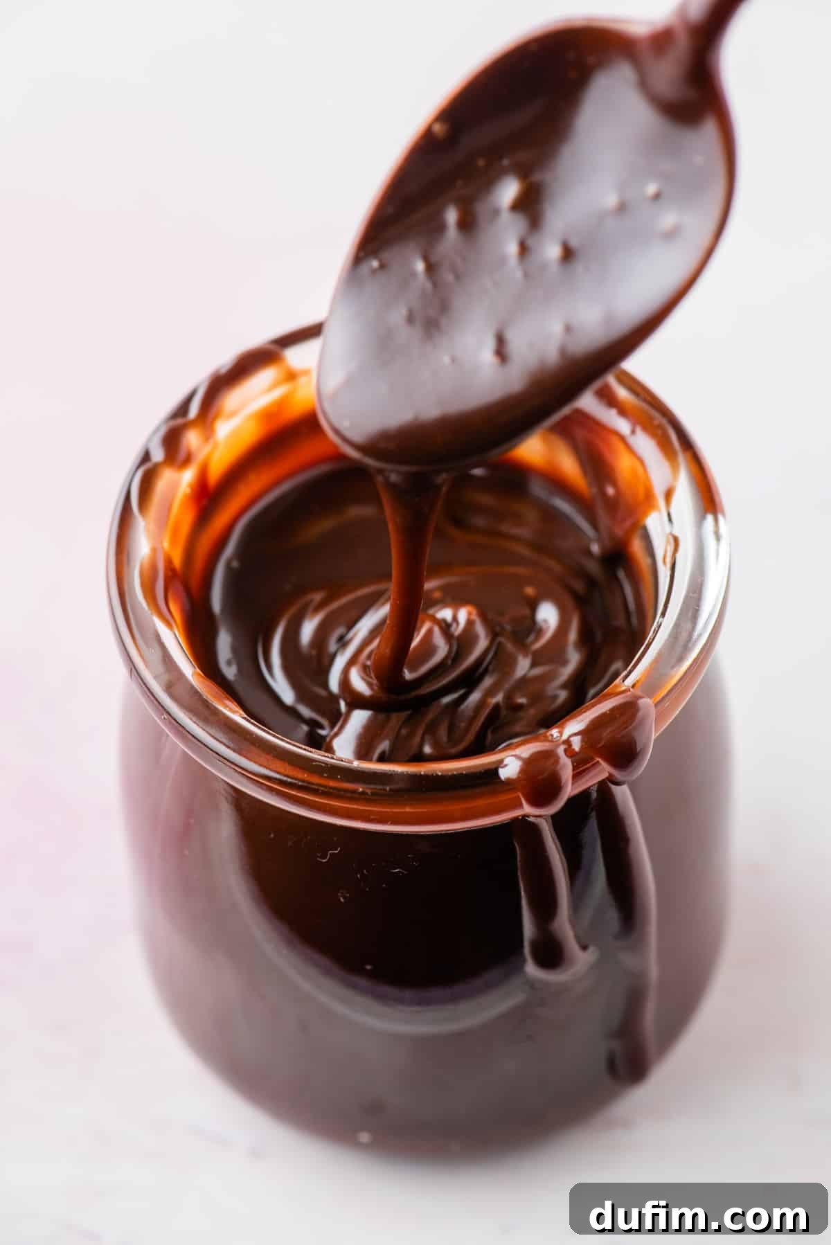 a jar of hot fudge sauce with a spoon above it with a drizzle of hot fudge sauce dripping down