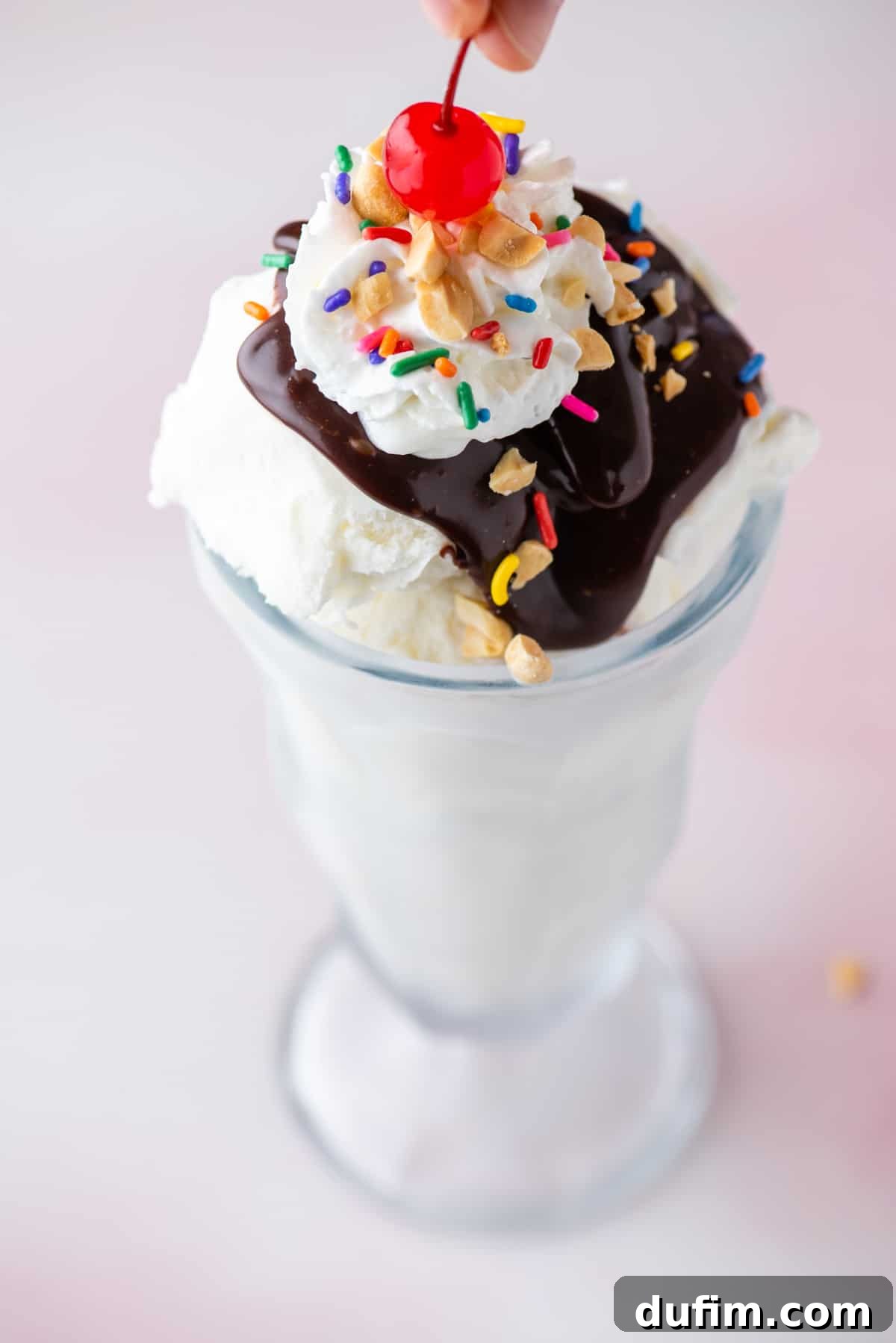 a hot fudge sundae in a tall glass topped with sprinkles, peanuts and cool whip with a cherry being placed on top