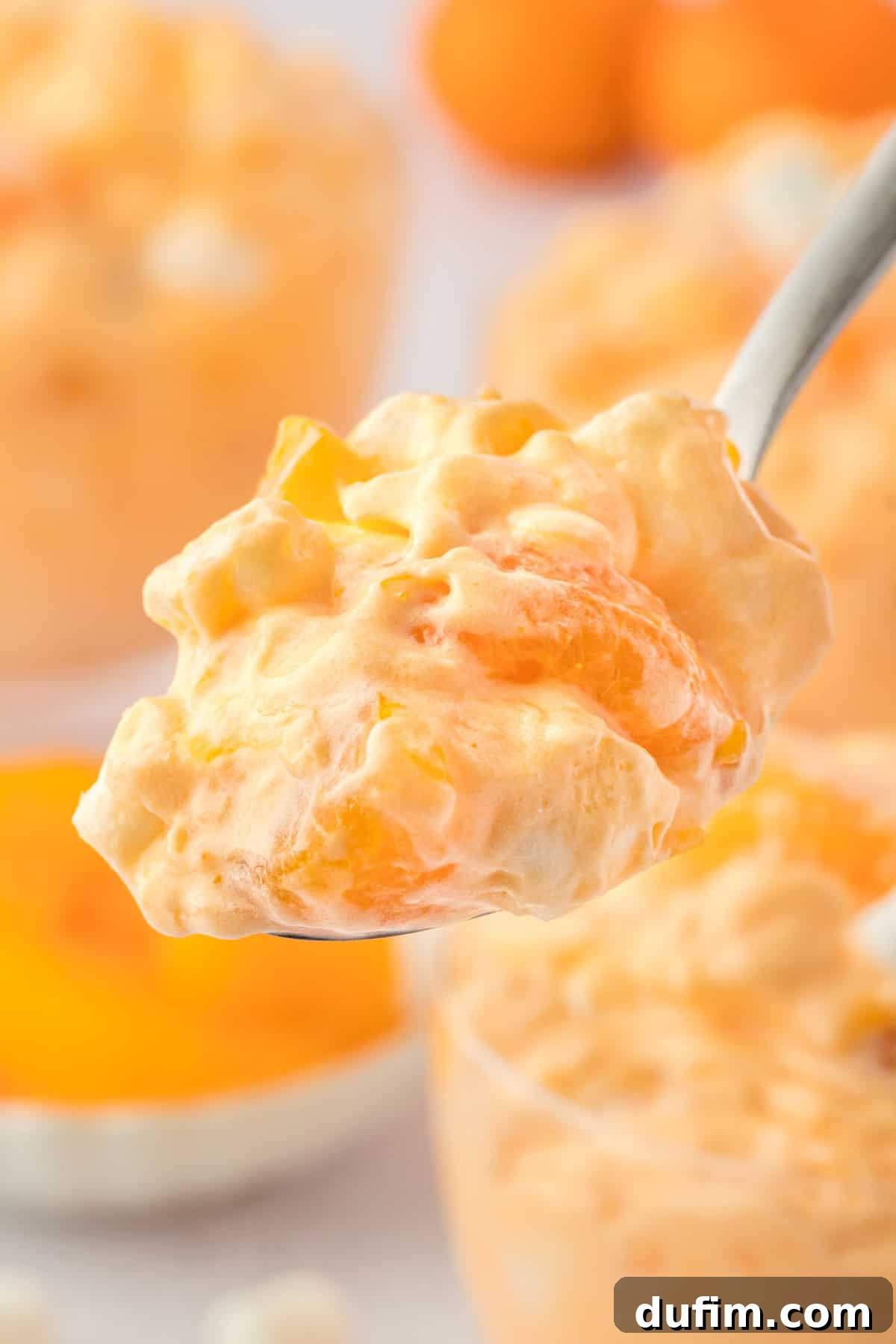 A close-up shot of a spoon laden with orange fluff salad, showcasing its creamy, airy texture and visible fruit pieces and marshmallows. In the softly blurred background, a larger bowl of the vibrant orange fluff and a separate bowl of fresh mandarin oranges are visible, highlighting the fresh and appealing nature of the ingredients.