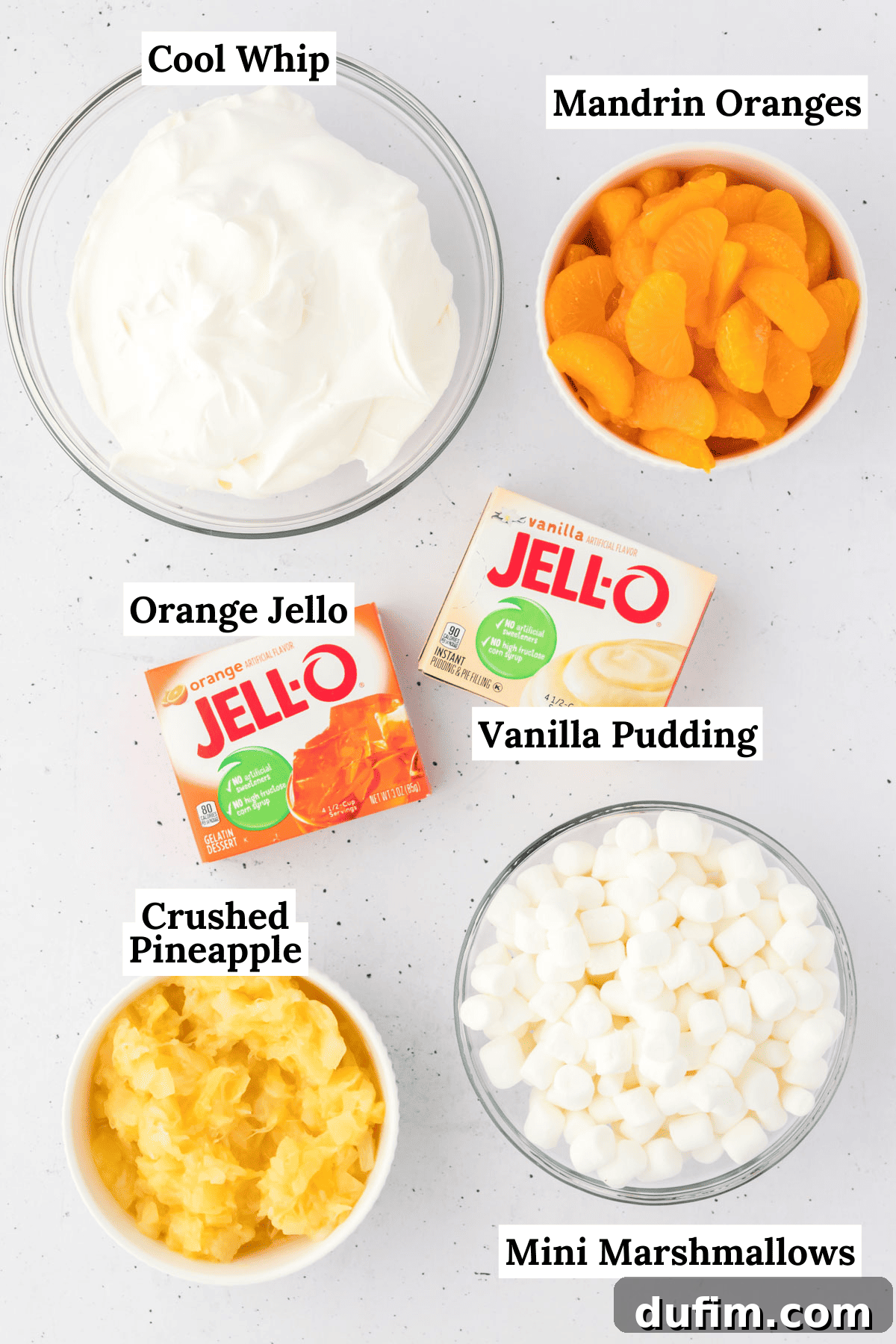 All the essential ingredients for Orange Fluff Salad are artfully arranged on a clean kitchen counter: a bright orange jello mix box, a vanilla pudding mix box, a clear bowl overflowing with fluffy mini marshmallows, a bowl of juicy crushed pineapple, and a container of creamy cool whip. This flat lay photography highlights the simplicity and readiness of the components, making preparation seem effortless.