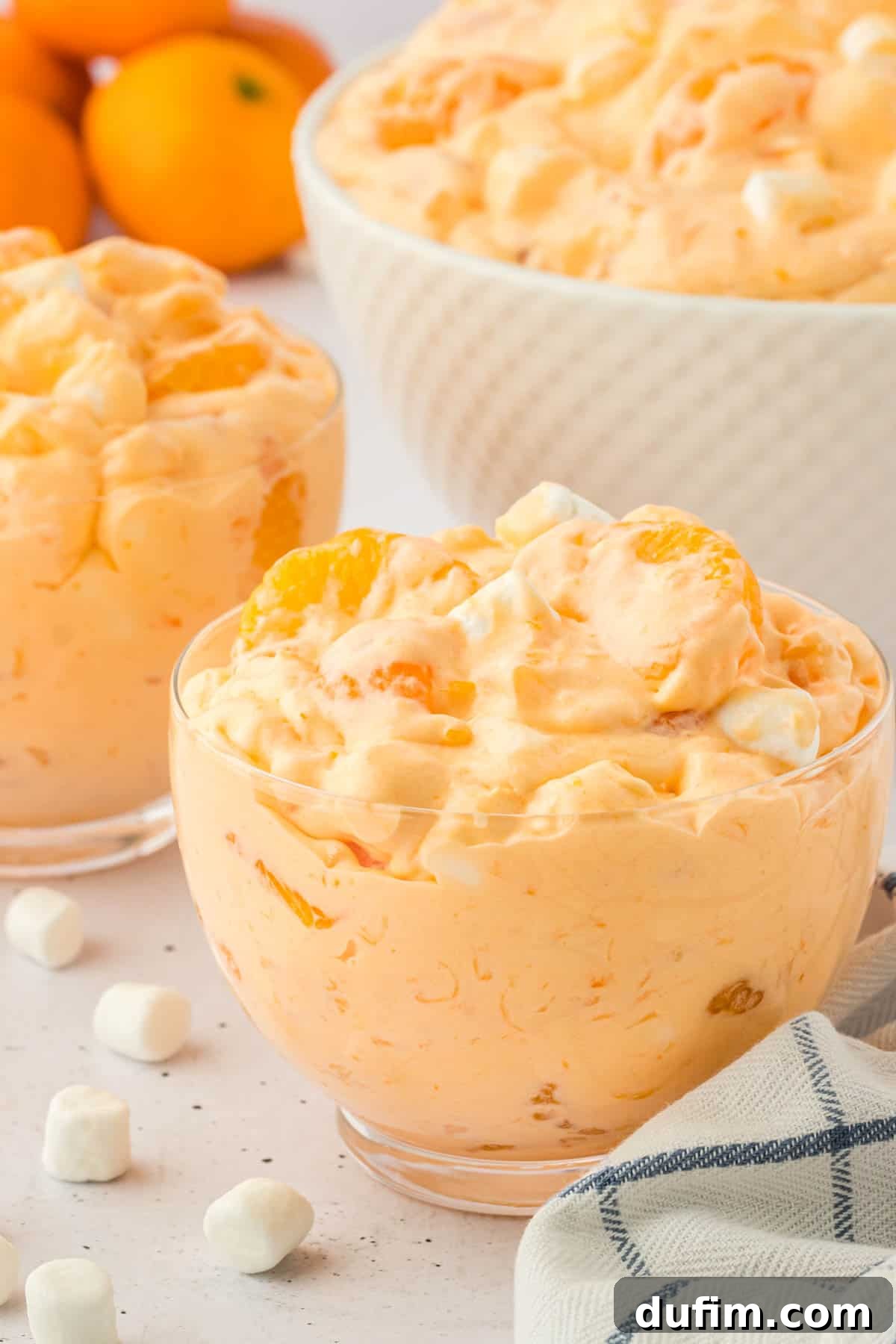 Three beautifully portioned bowls of fluffy orange fluff salad, each invitingly arranged. They are artfully set on a clean surface, surrounded by scattered mini marshmallows and a neatly folded blue and white kitchen towel, creating a charming and homey scene that highlights the dish's appeal.