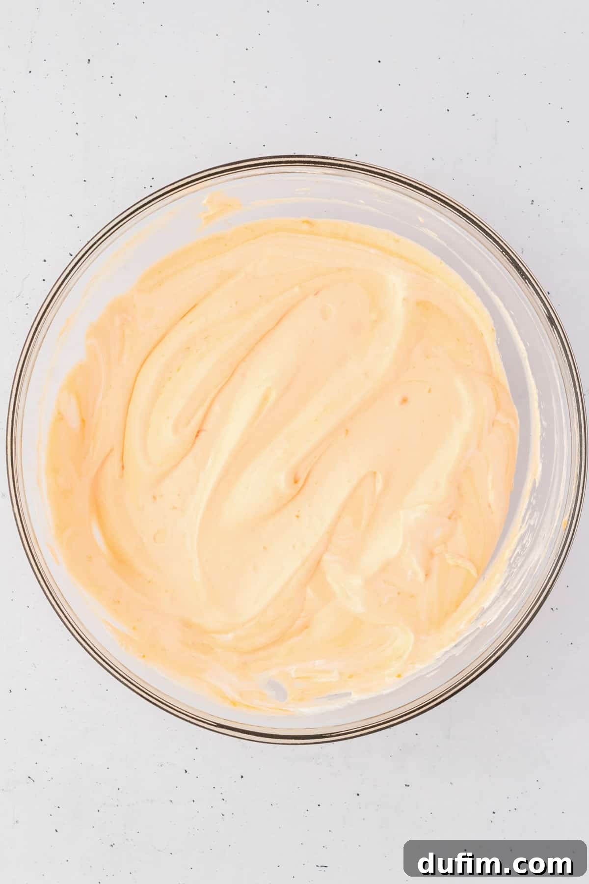 A pristine glass bowl containing the creamy, light orange base mixture for orange fluff salad, captured before the addition of marshmallows and fruit. The smooth, whipped, and partially set texture is evident, showcasing its readiness for the final flavorful additions.