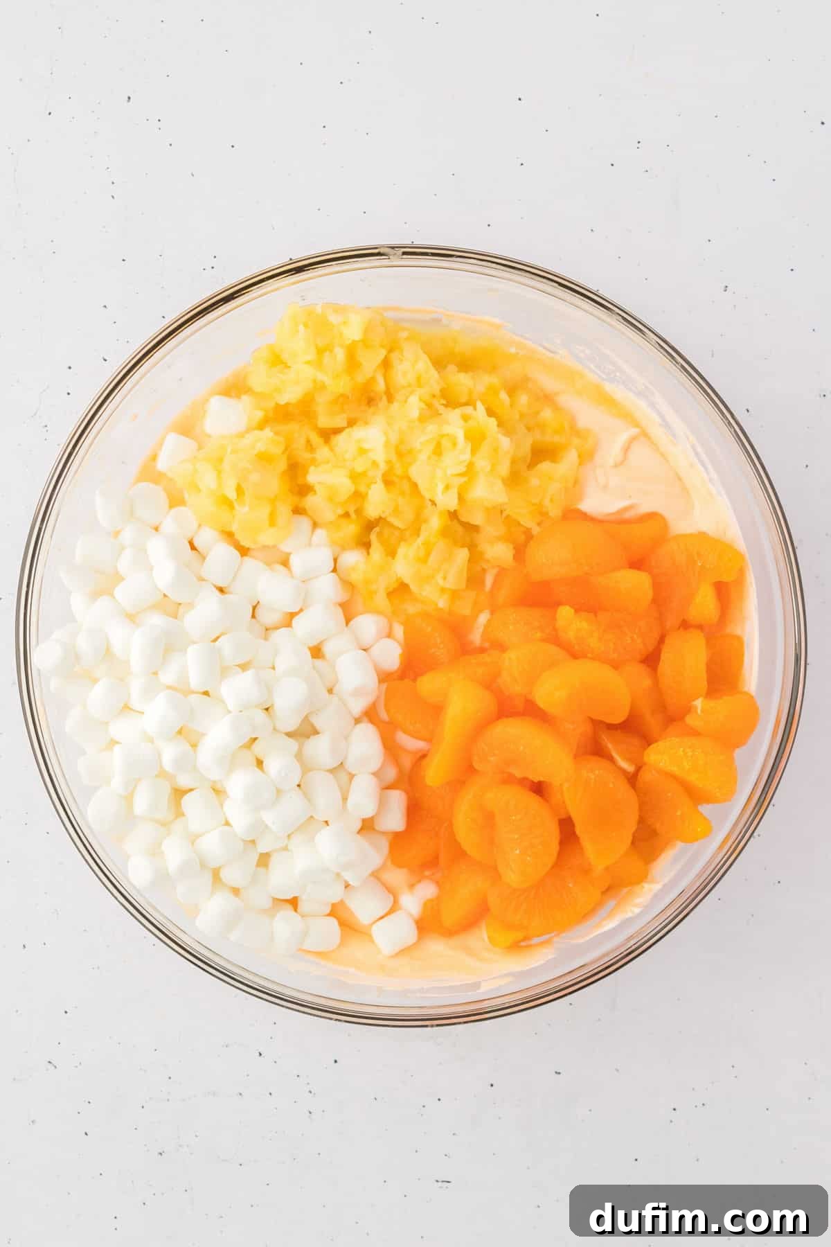 The creamy, light orange base for fluff salad, now generously topped with vibrant mini marshmallows, glistening crushed pineapple, and bright mandarin oranges, all awaiting their gentle fold into the mixture. This image captures the dish just before its final combination, highlighting the fresh and colorful components.
