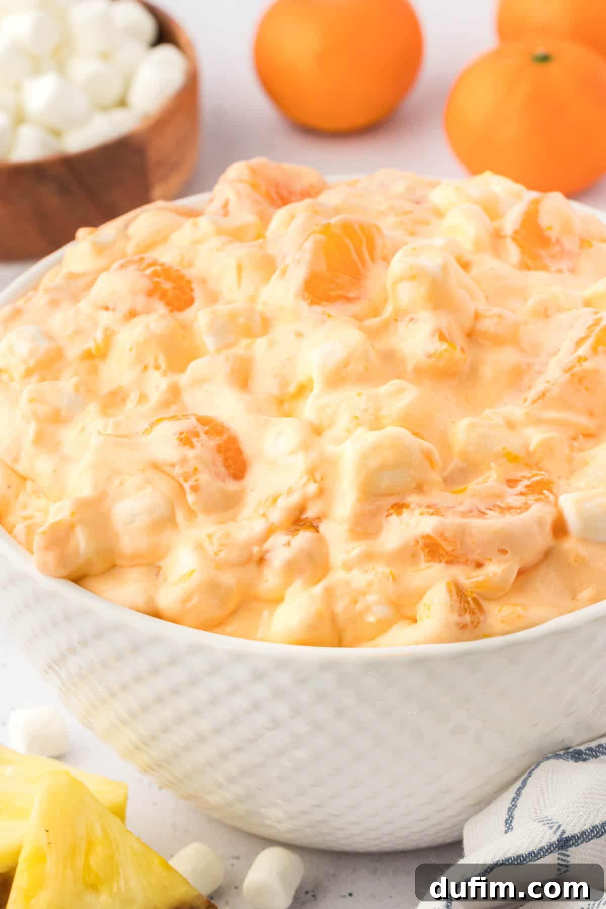 A beautifully presented bowl of vibrant orange fluff salad, its rich color and fluffy texture instantly appealing. It is artfully surrounded by a small bowl of extra mini marshmallows, whole and sliced fresh mandarin oranges, elegant pineapple wedges, and a stylish blue and white kitchen towel. This arrangement thoughtfully highlights the fresh and sweet components of the dessert, inviting viewers to savor its delightful taste.