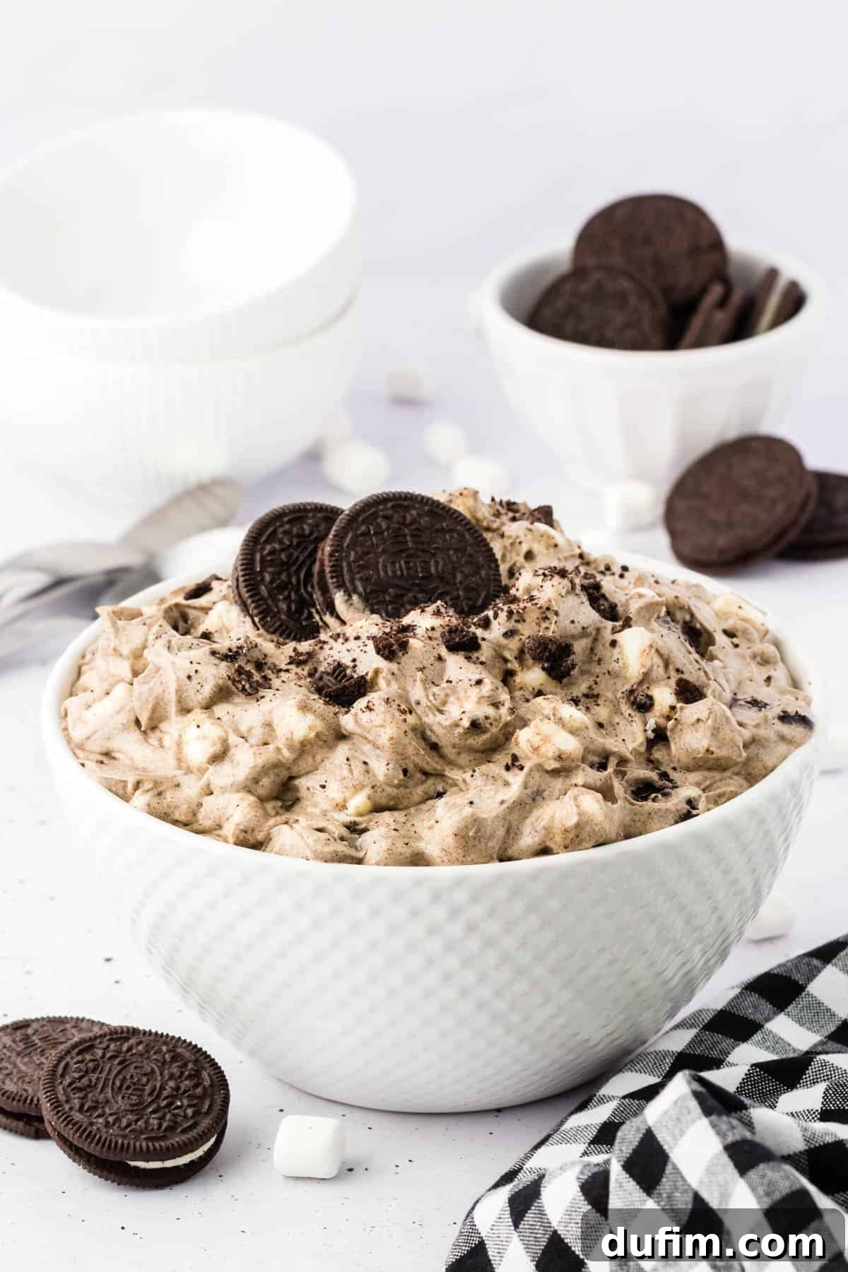 No-Fuss Oreo Fluff 2 A generous serving of Oreo fluff in a pristine white bowl, surrounded by whole Oreo cookies, fluffy mini marshmallows, and a classic black and white checkered kitchen towel, hinting at its homemade goodness.