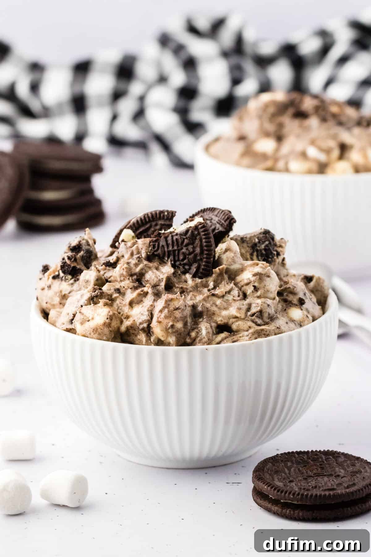 No-Fuss Oreo Fluff 12 A beautifully presented bowl of Oreo fluff, set against a backdrop of whole Oreo cookies, mini marshmallows, and a classic black and white checkered kitchen towel, showcasing its inviting texture.