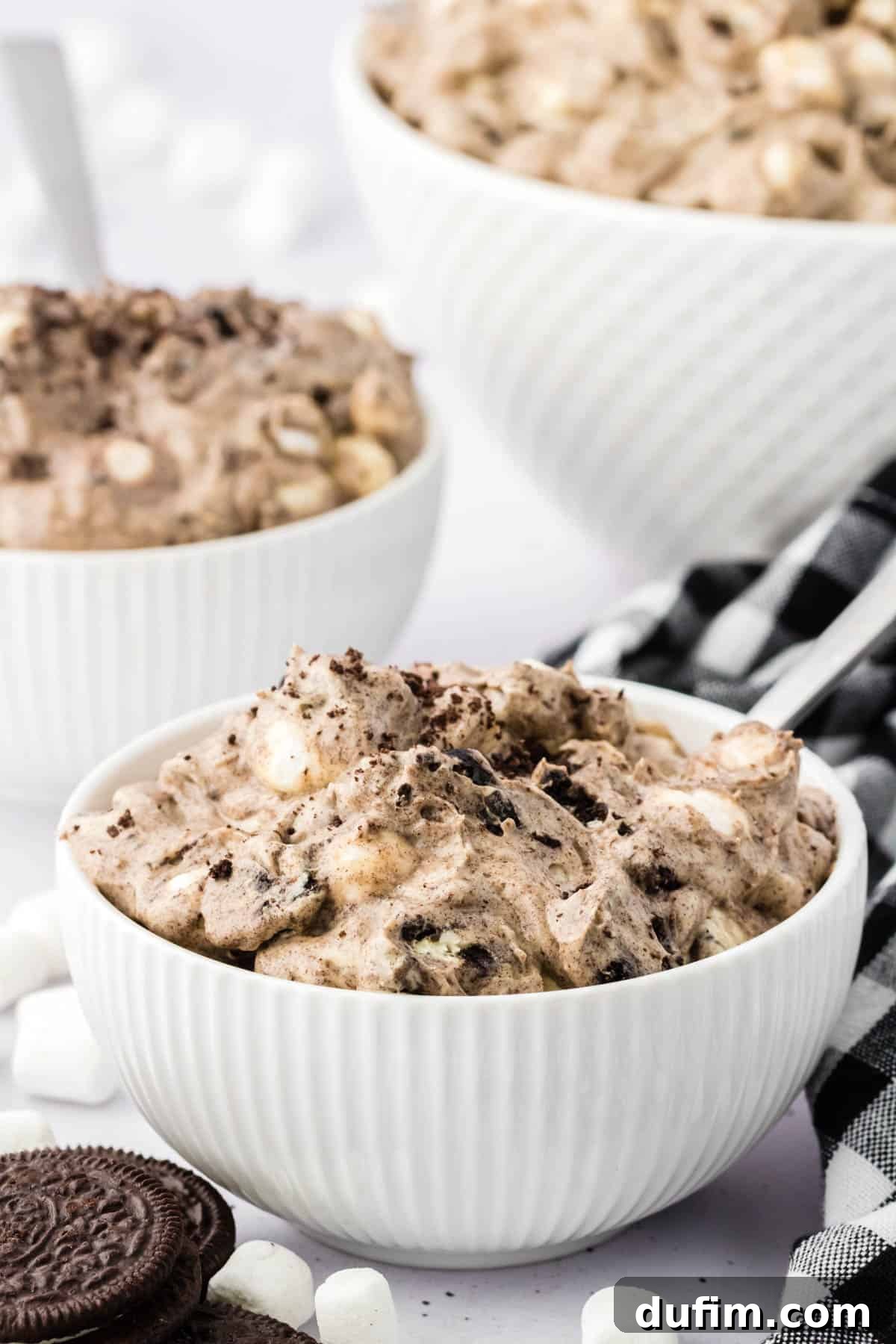 No-Fuss Oreo Fluff 3 Three tempting bowls of Oreo fluff, perfectly arranged and ready to be served, complemented by whole Oreos, mini marshmallows, and a stylish black and white checkered kitchen towel, showcasing its visual appeal.