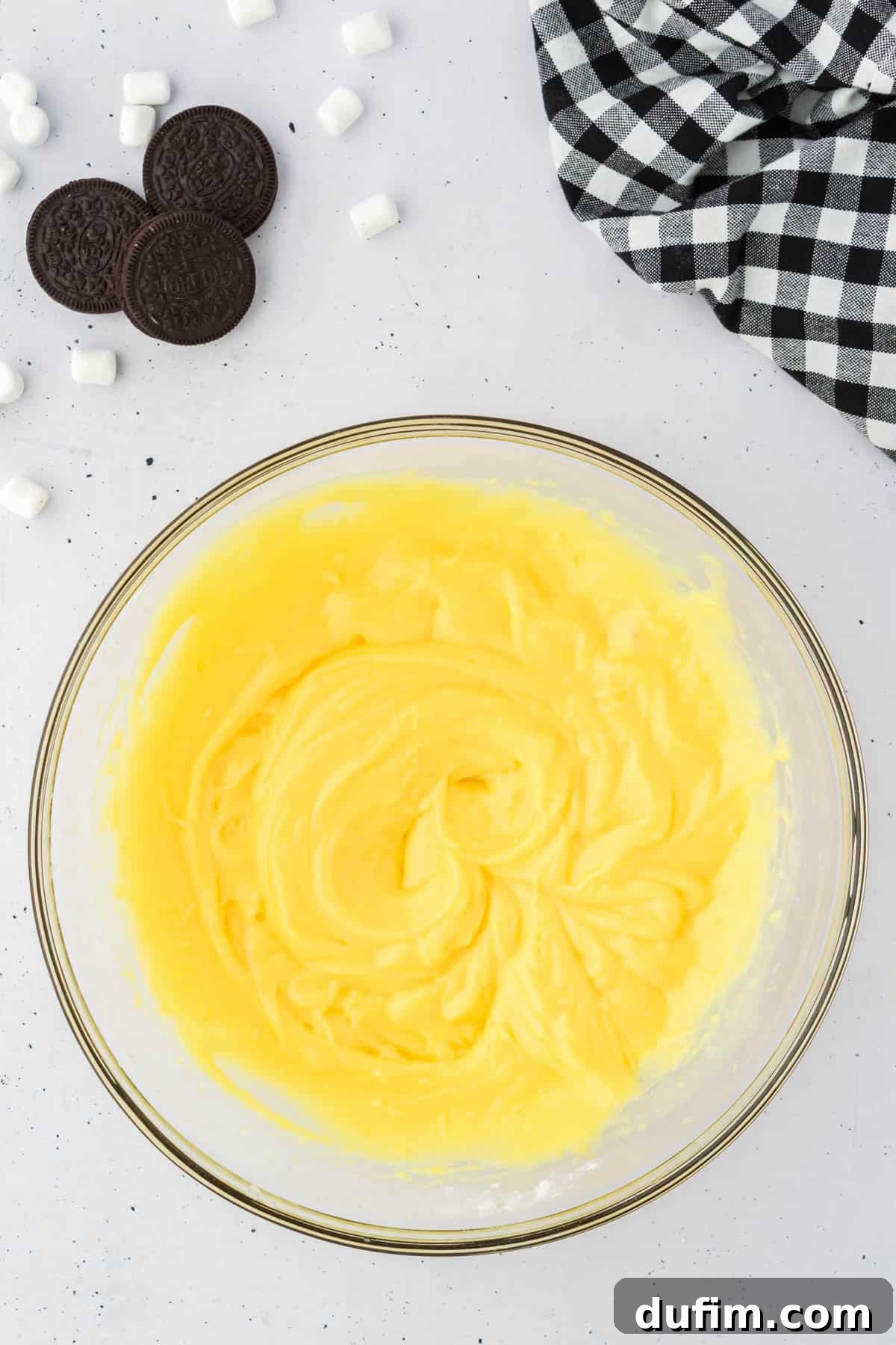 No-Fuss Oreo Fluff 5 A bowl filled with smoothly mixed vanilla pudding, ready for the next steps, surrounded by whole Oreos, mini marshmallows, and a black and white checkered kitchen towel.